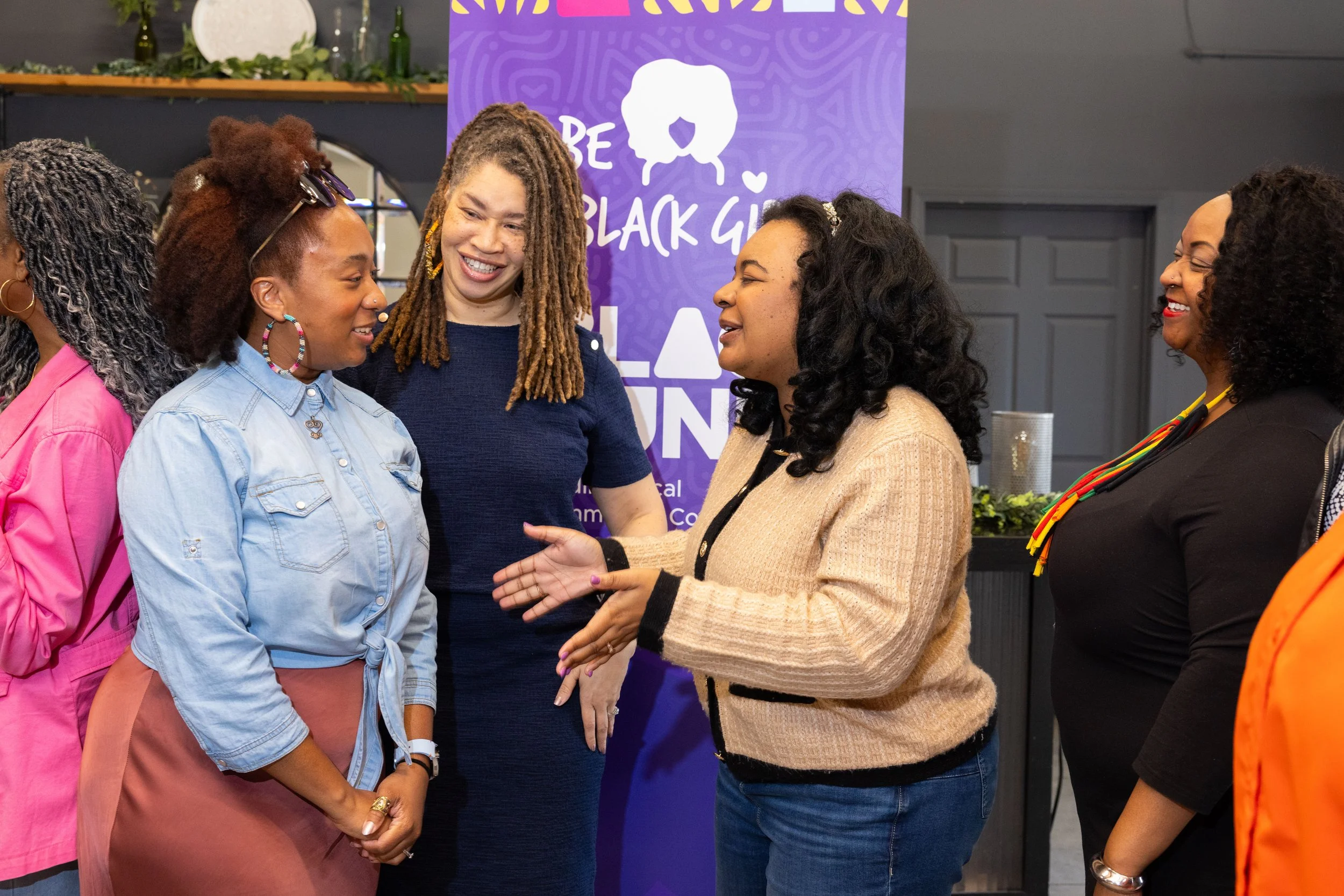 A diverse group of women engaged in conversation at an indoor event. They are smiling and appear to be enjoying each other's company. In the background, there is a purple sign with white text and graphics.