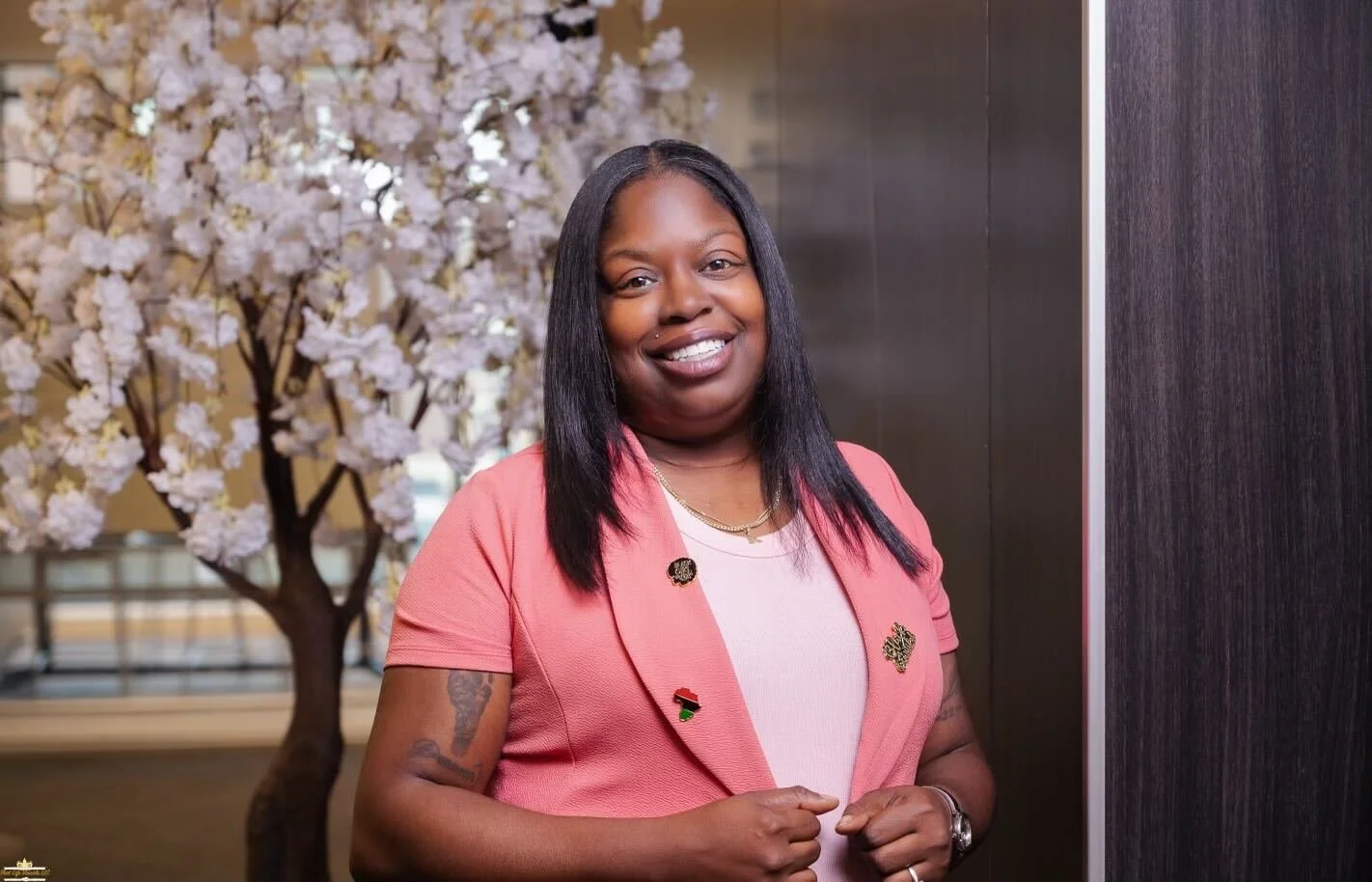Smiling woman with shoulder-length black hair, wearing a pink blazer with pins, standing indoors near a dark wall, with a decorative pink cherry blossom tree in the background.