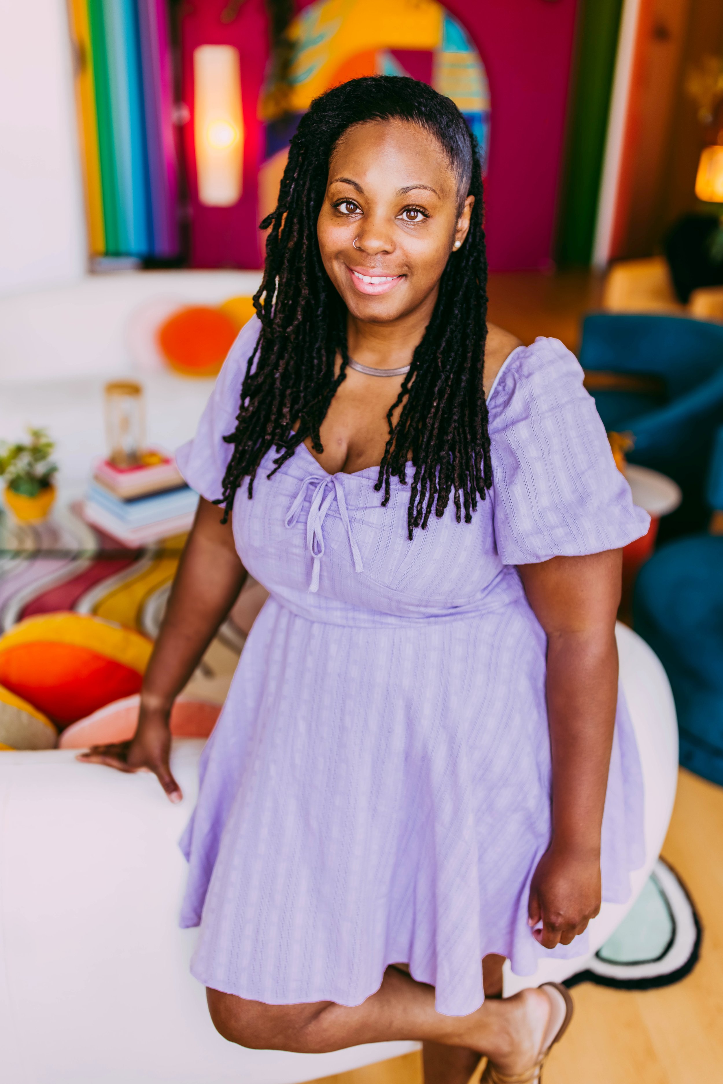 A woman with long dreadlocks wearing a purple dress and smiling at the camera in an art studio with colorful artwork and rainbow decorations in the background.