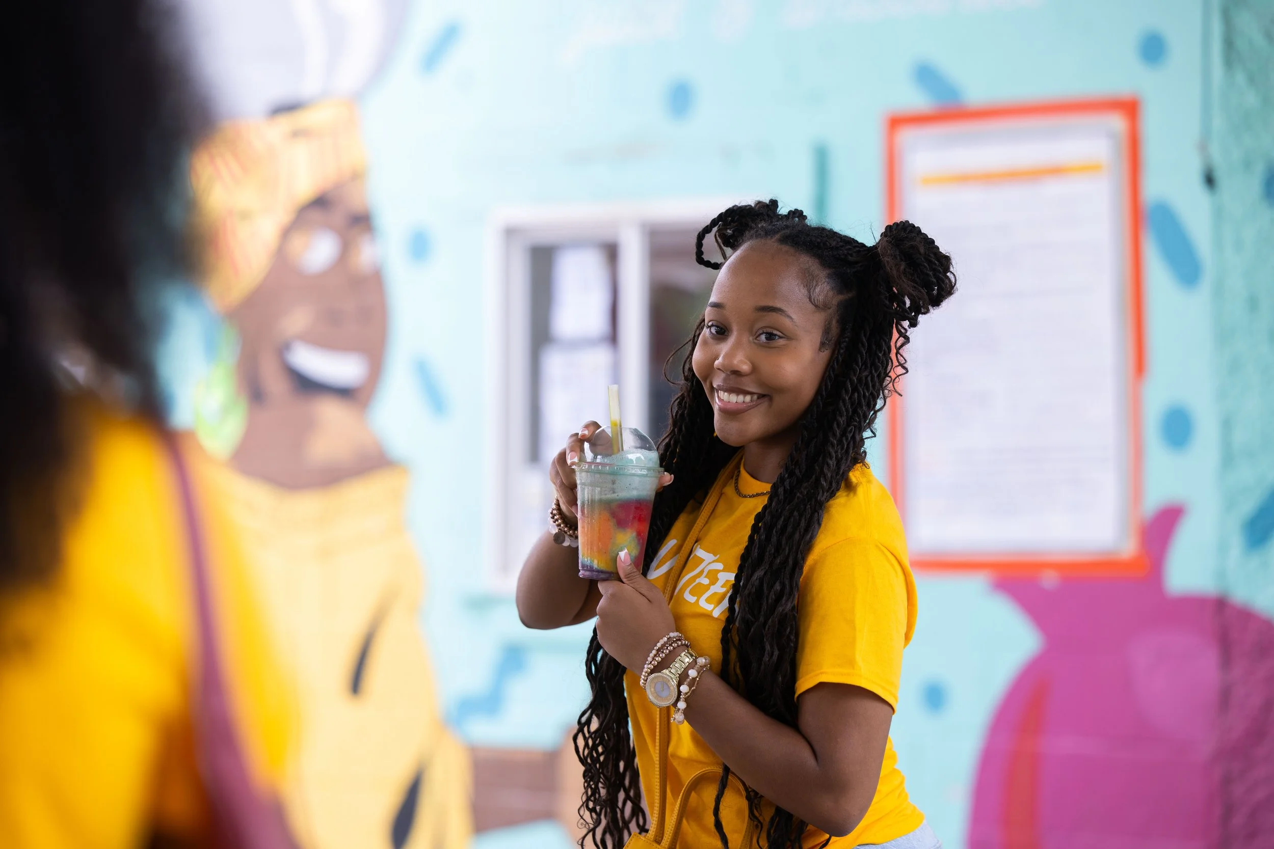 A young girl with long braided hair, wearing a yellow shirt, smiling and holding a colorful drink, standing in front of a bright blue wall with colorful decorations.