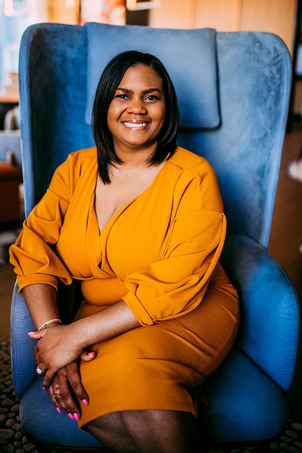 A woman with shoulder-length black hair and a bright smile is sitting in a high-backed, blue velvet armchair, wearing a mustard yellow dress, in an indoor setting.