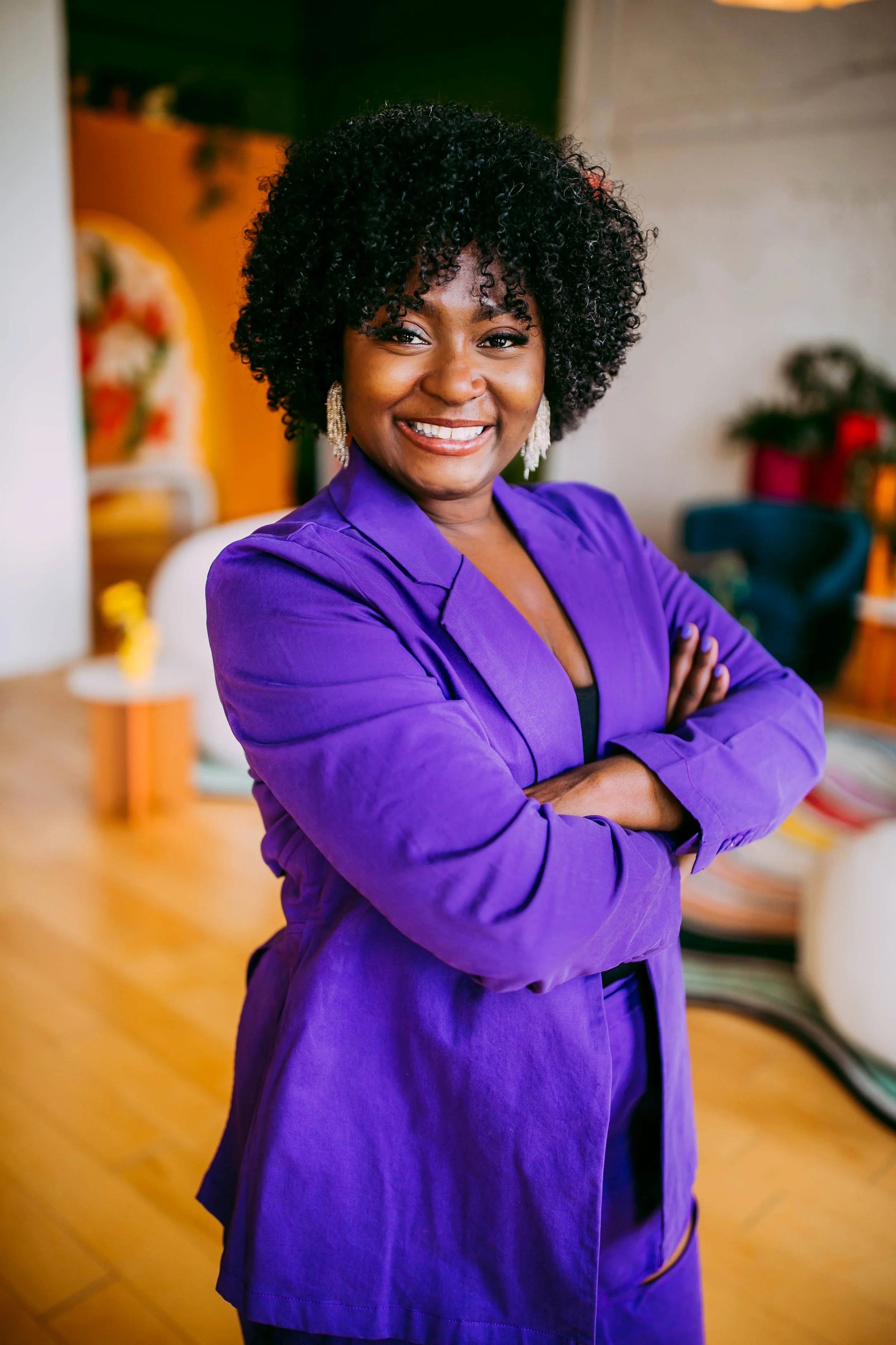A woman with curly black hair and earrings, smiling, wearing a purple blazer with crossing arms, standing indoors with blurred colorful background.