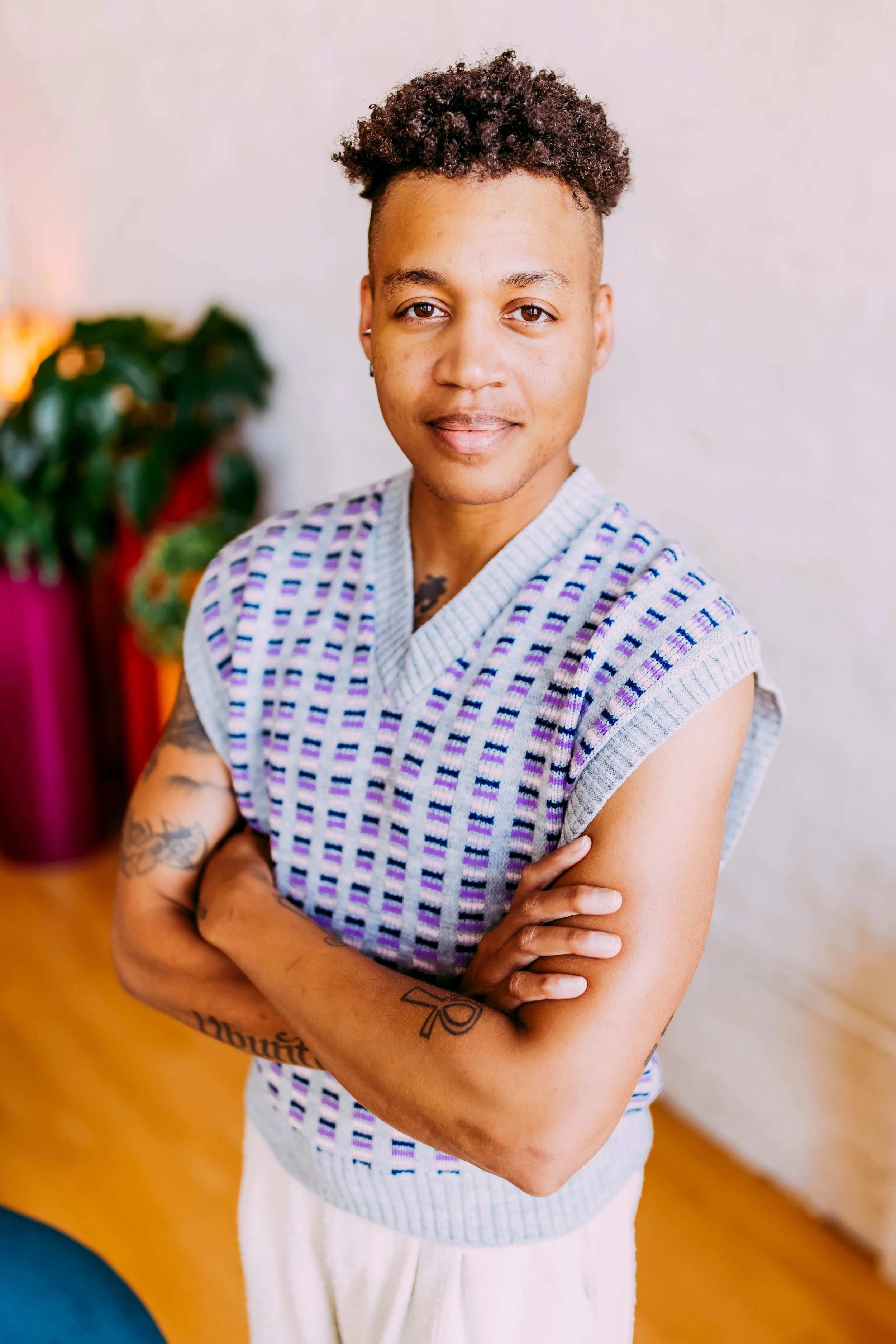 A person with short curly hair and tattoos on their arms, wearing a patterned sleeveless shirt and white pants, standing indoors with arms crossed, smiling at the camera, with a blurred plant and colorful background.