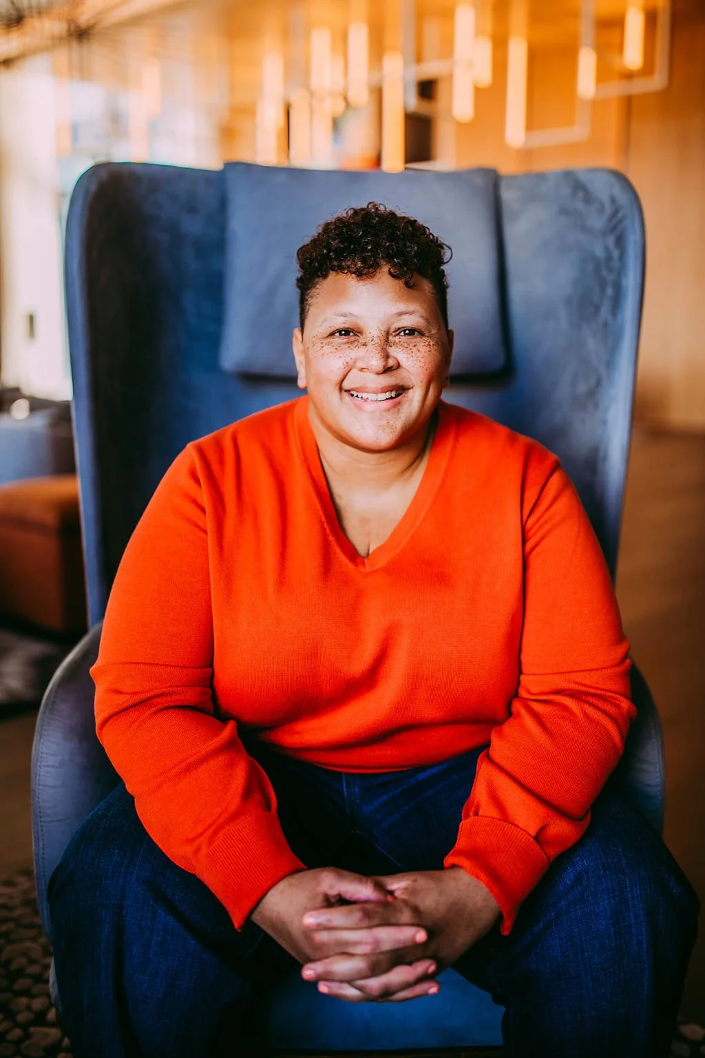 A woman with short curly hair and freckles, smiling, sitting in a modern chair in a wooden interior space, wearing a bright orange sweater and dark jeans.