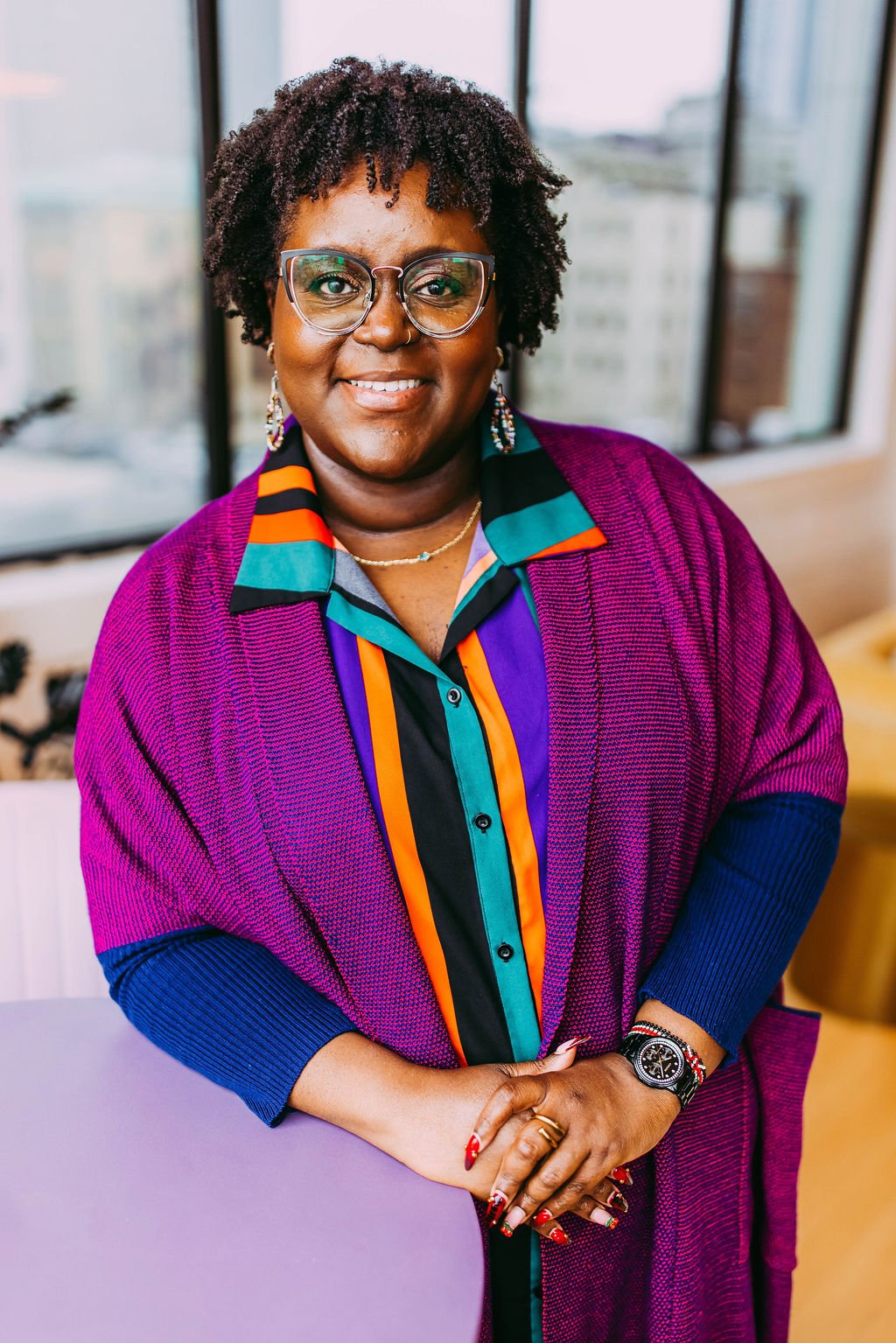 A smiling woman with glasses, short curly hair, and earrings, wearing a colorful patterned shirt and a purple cardigan, standing indoors near large windows.