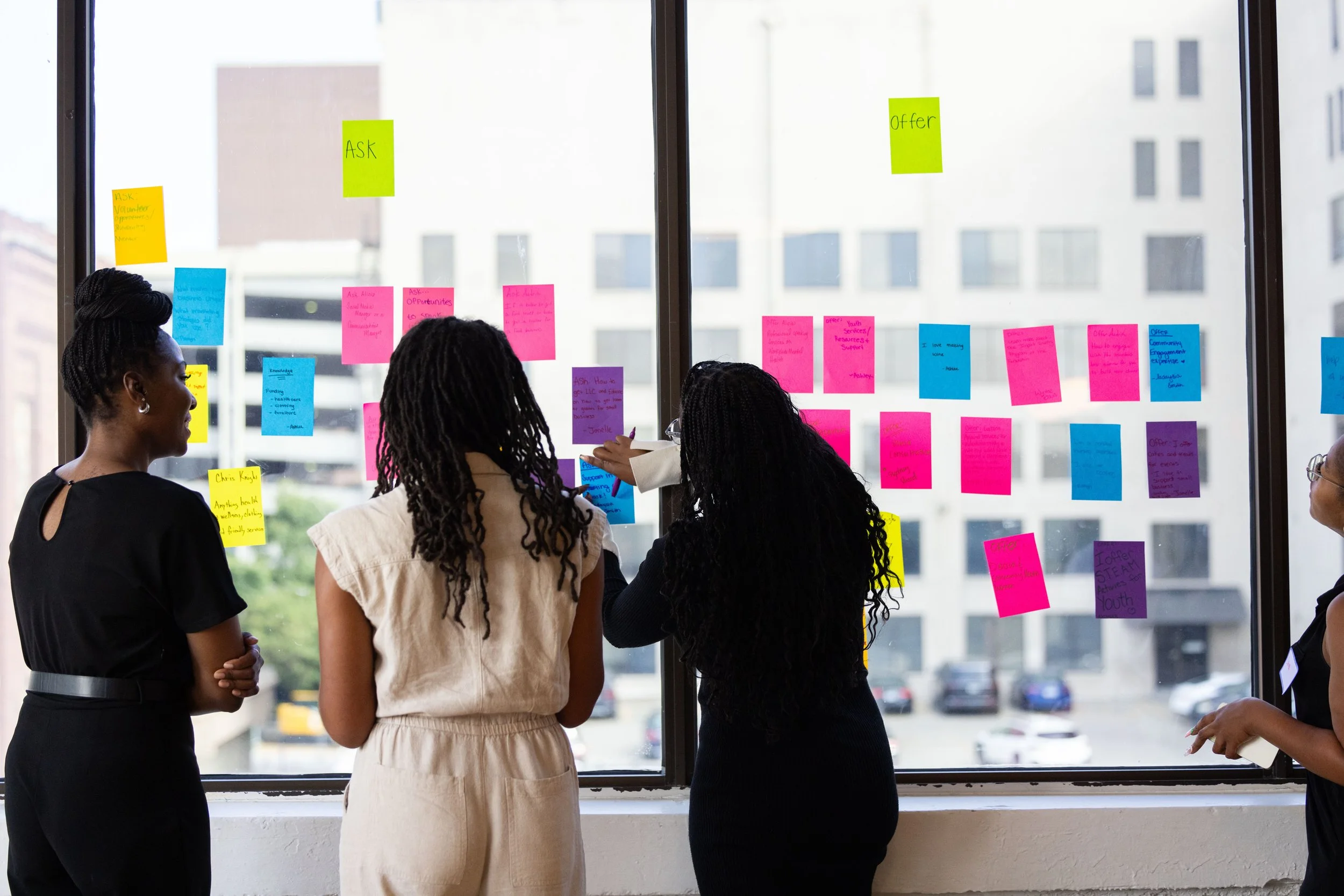 Four women standing by a large window covered with colorful sticky notes containing handwritten text.