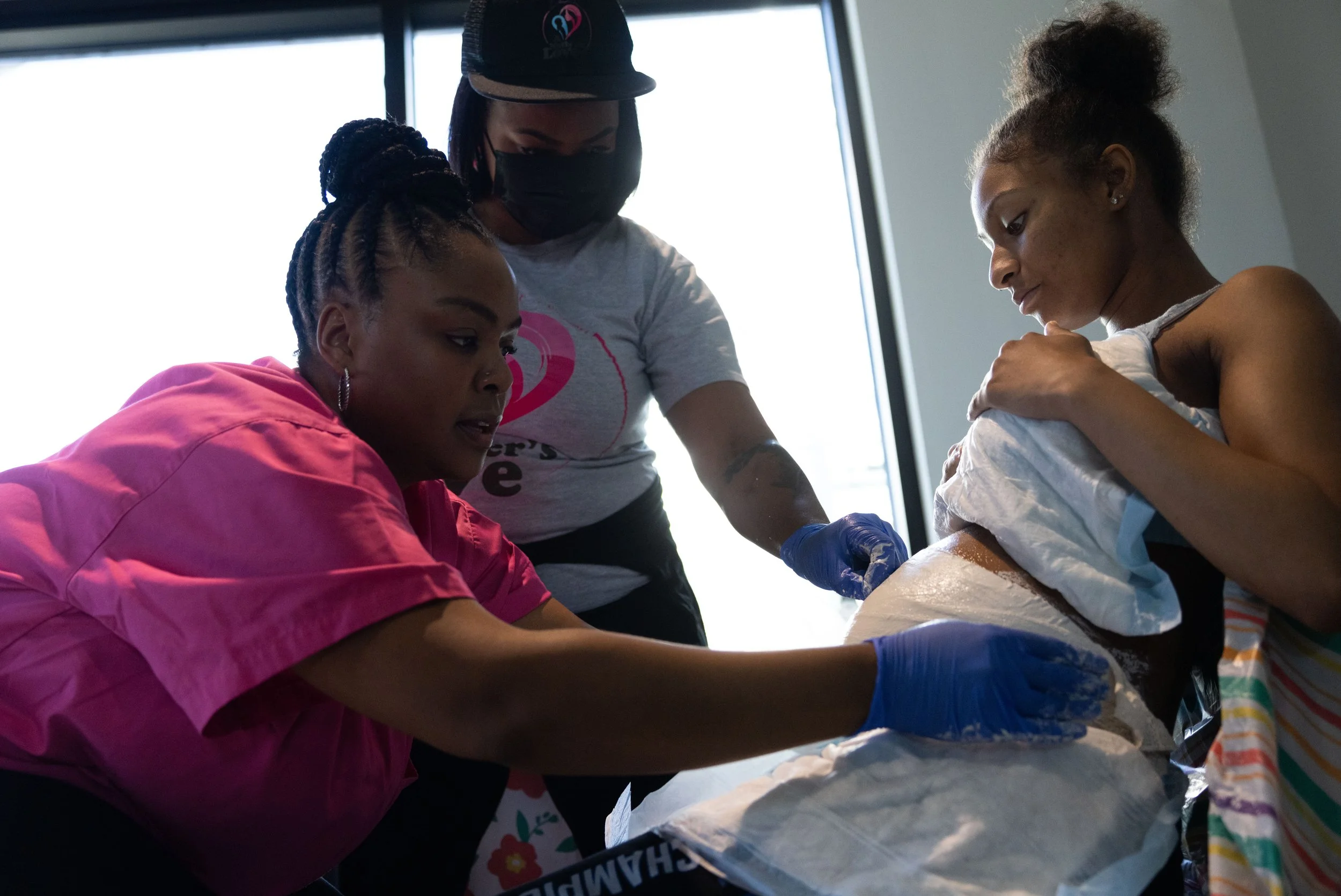 A medical professional and two women in a clinical setting. The woman on the right is lying on a table, covered with a blanket, and is being prepared for a medical procedure. The woman on the left, wearing a pink medical smock, is assisting and holding the woman's arm. The professional, wearing a gray shirt, a cap, a face mask, and blue gloves, is tending to the woman on the table.