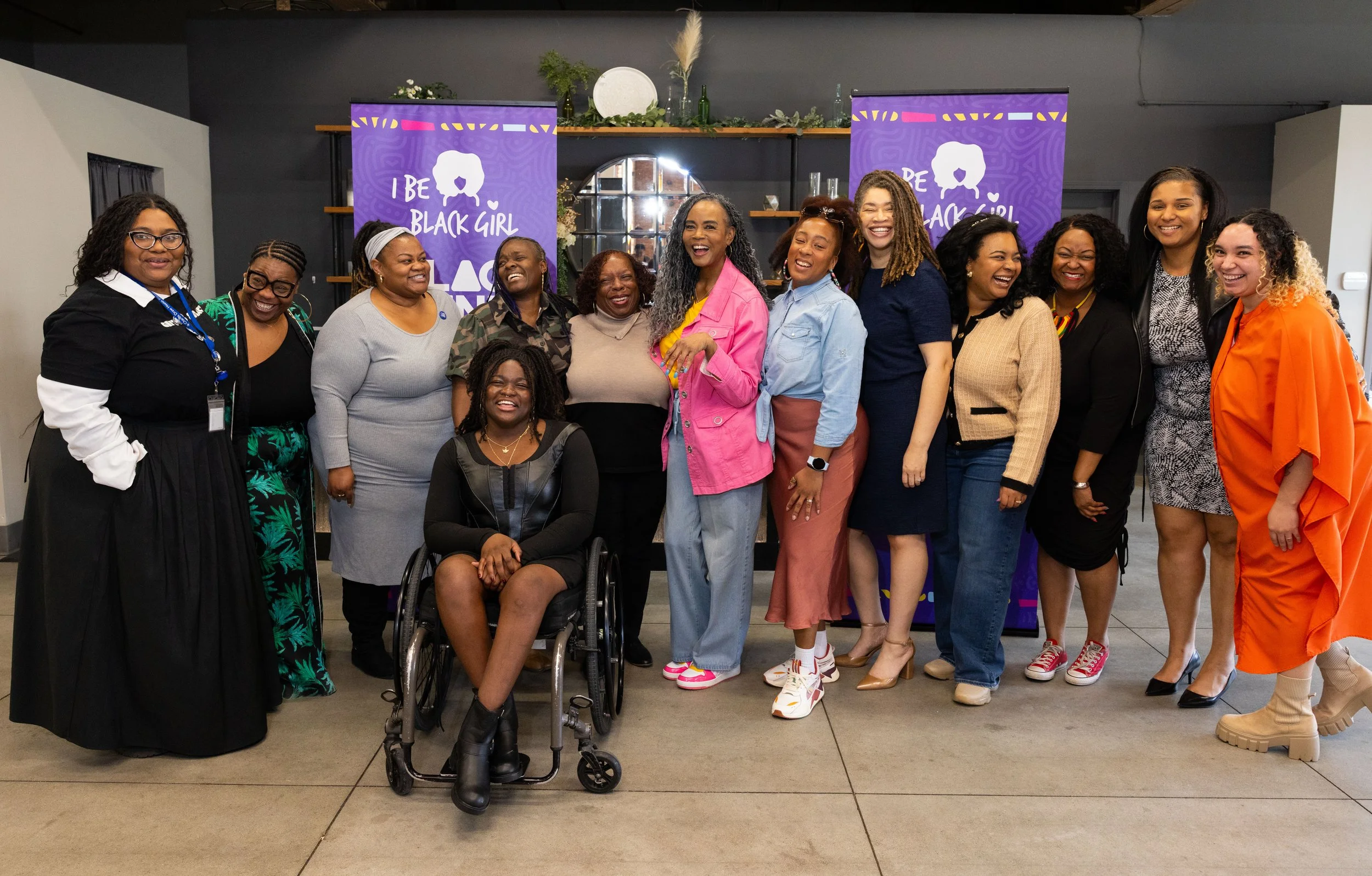 Group of diverse women smiling and laughing in an indoor space with purple banners that say "I BE BLACK GIRL" behind them.