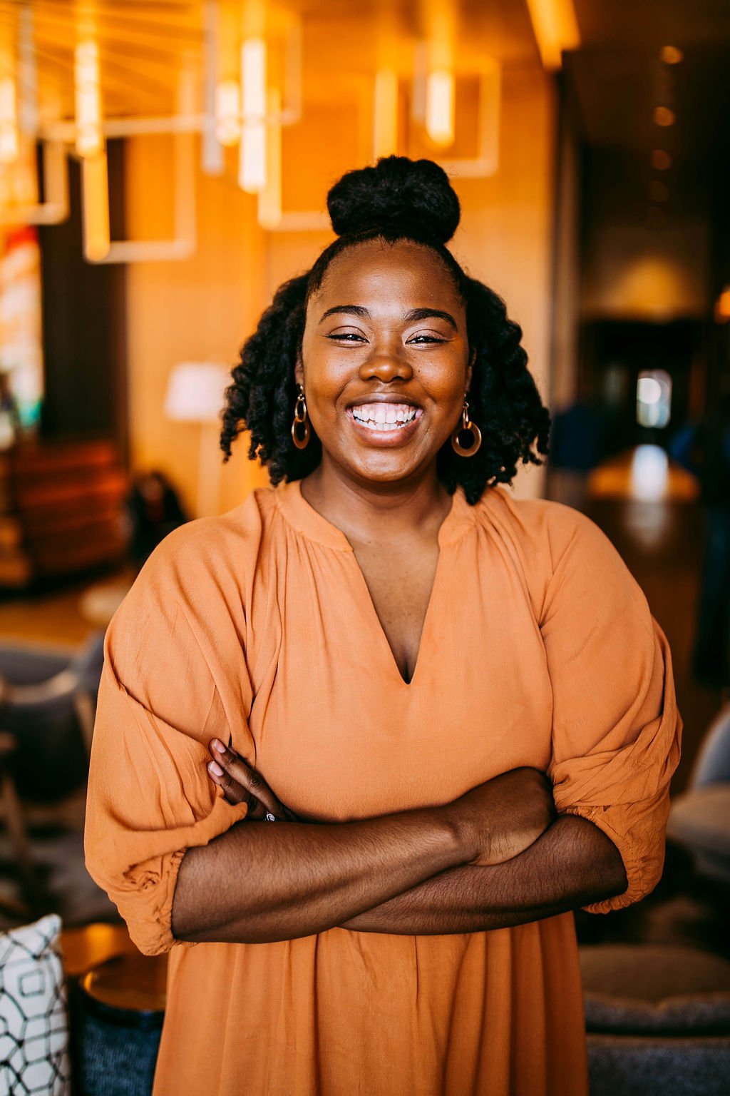 A woman smiling with arms crossed in a warmly lit room, wearing gold hoop earrings and an orange dress.