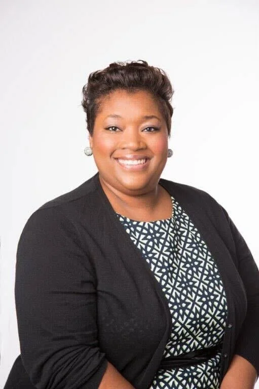 Portrait of a smiling Black woman with short curly hair, wearing a black blazer and a patterned blouse, against a white background.