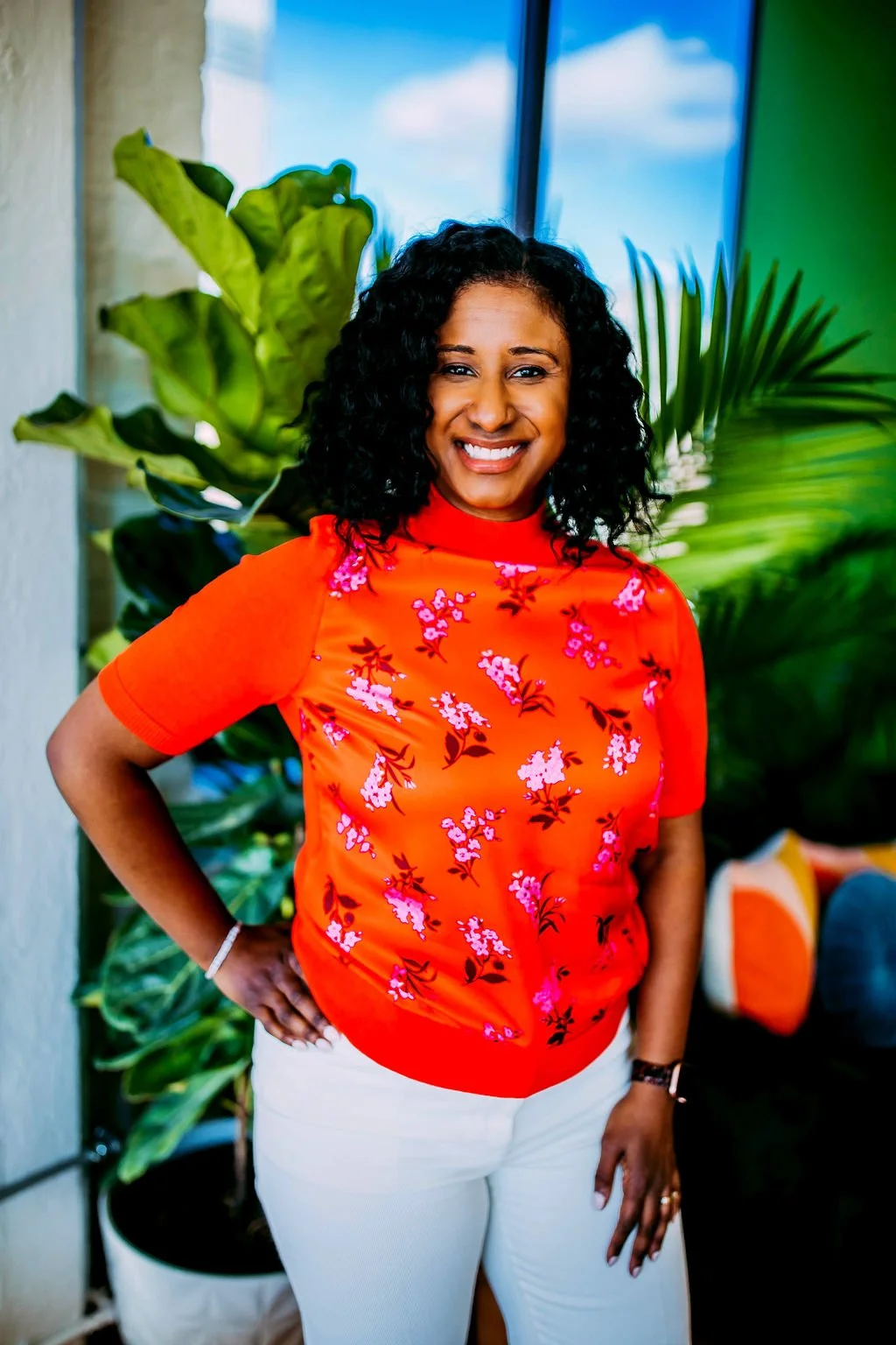 A woman with curly black hair smiling, wearing an orange floral blouse and white pants, standing indoors with green plants in the background.