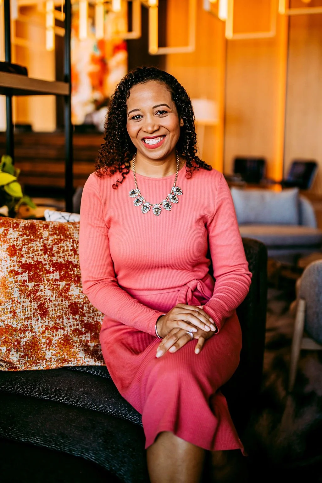 A woman with curly black hair wearing a pink dress and necklace, sitting on a sofa in a warmly lit modern interior.