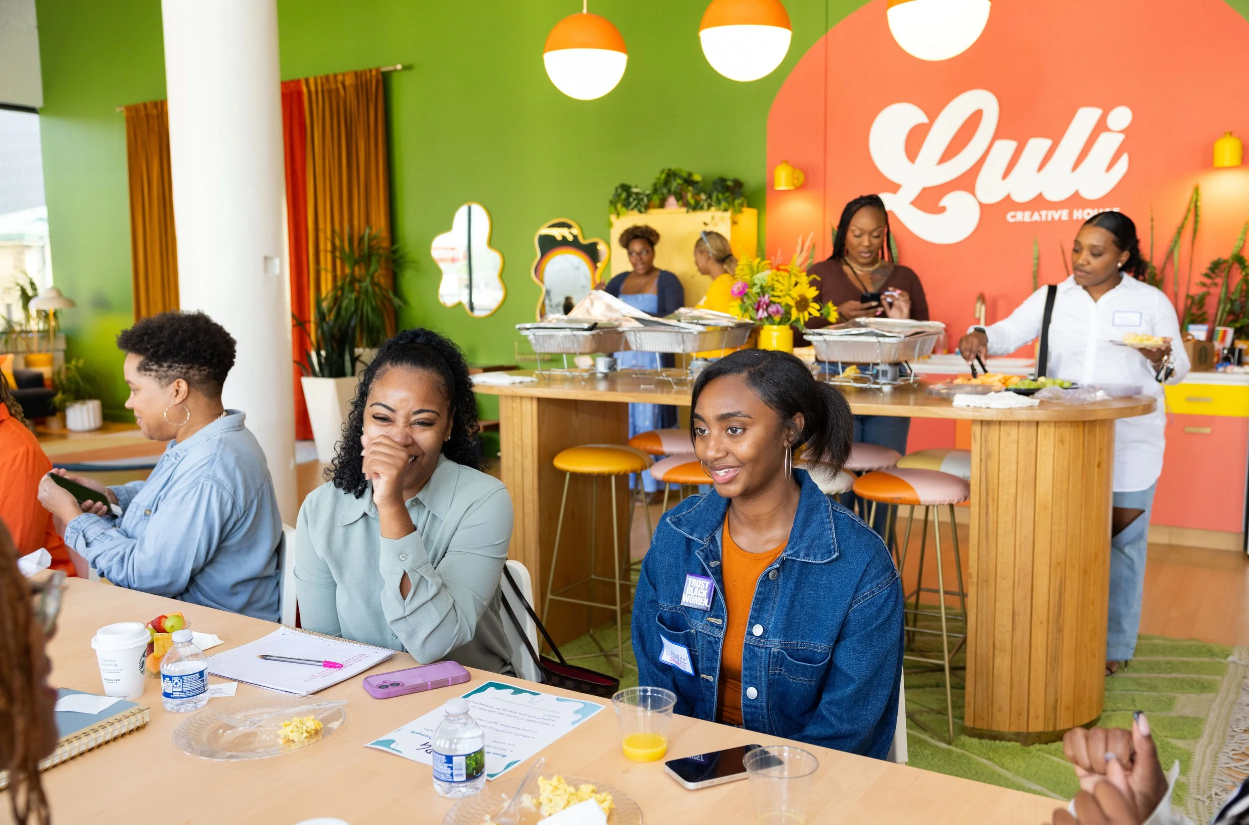 Women sitting at a table during a meeting or workshop in a colorful, vibrant room with decorative plants and a sign that reads 'Luli Creative House' on the wall.
