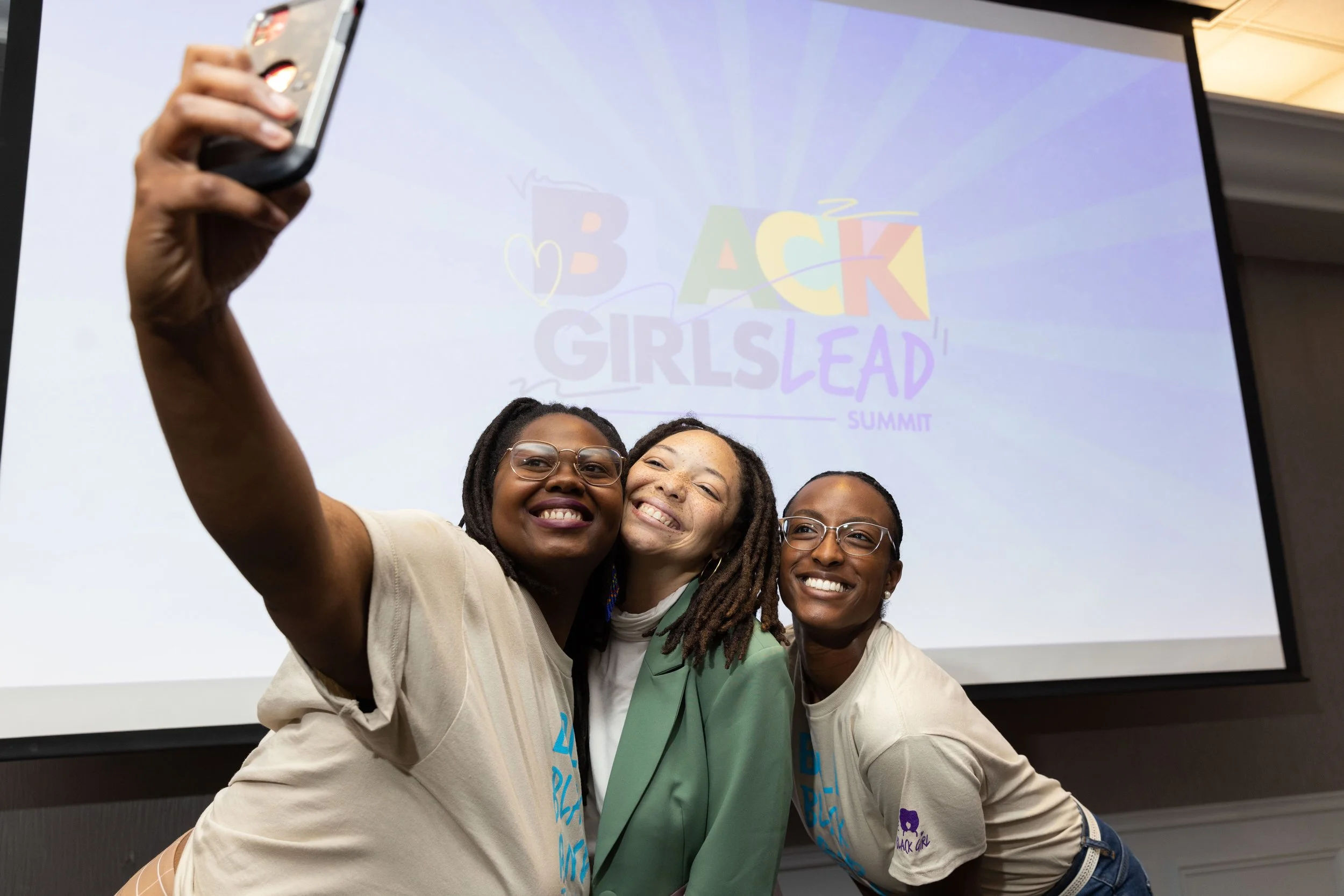Four women smiling and taking a selfie together at the Black Girls Lead Summit, with a screen displaying the event's logo in the background.