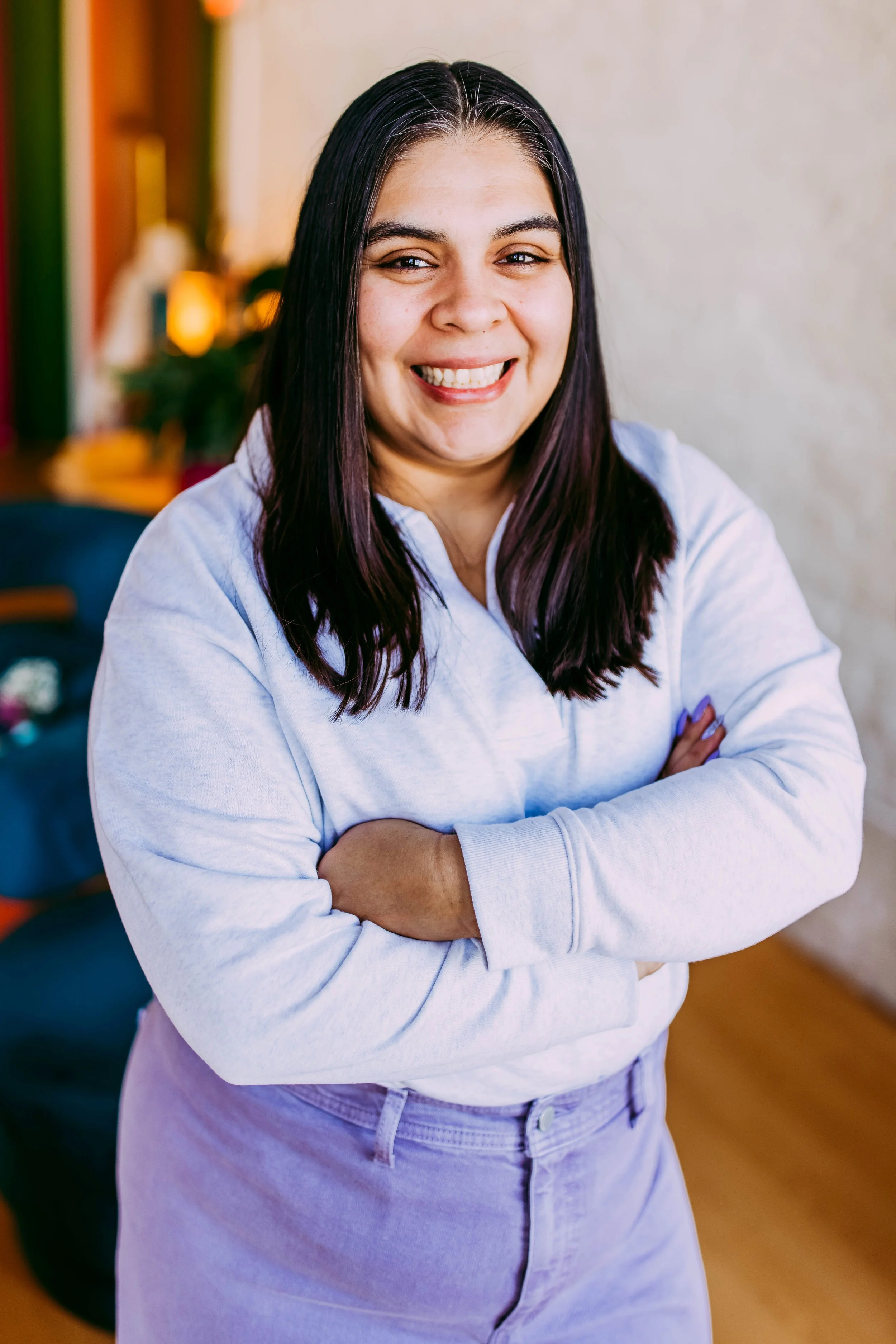 A smiling woman with dark hair and crossed arms, wearing a white sweater and light purple pants, in a cozy indoor setting.