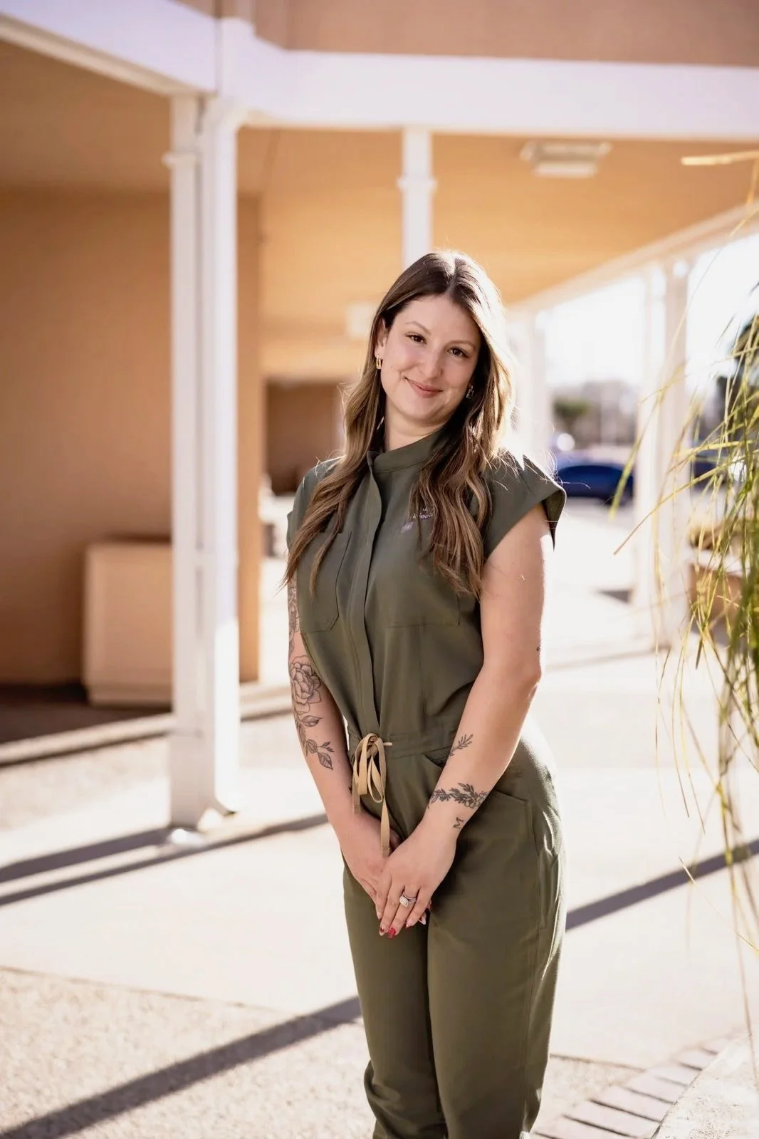 A woman standing outdoors, smiling, wearing an olive green jumpsuit, with tattoos on her arms, in front of a light-colored building on a sunny day.