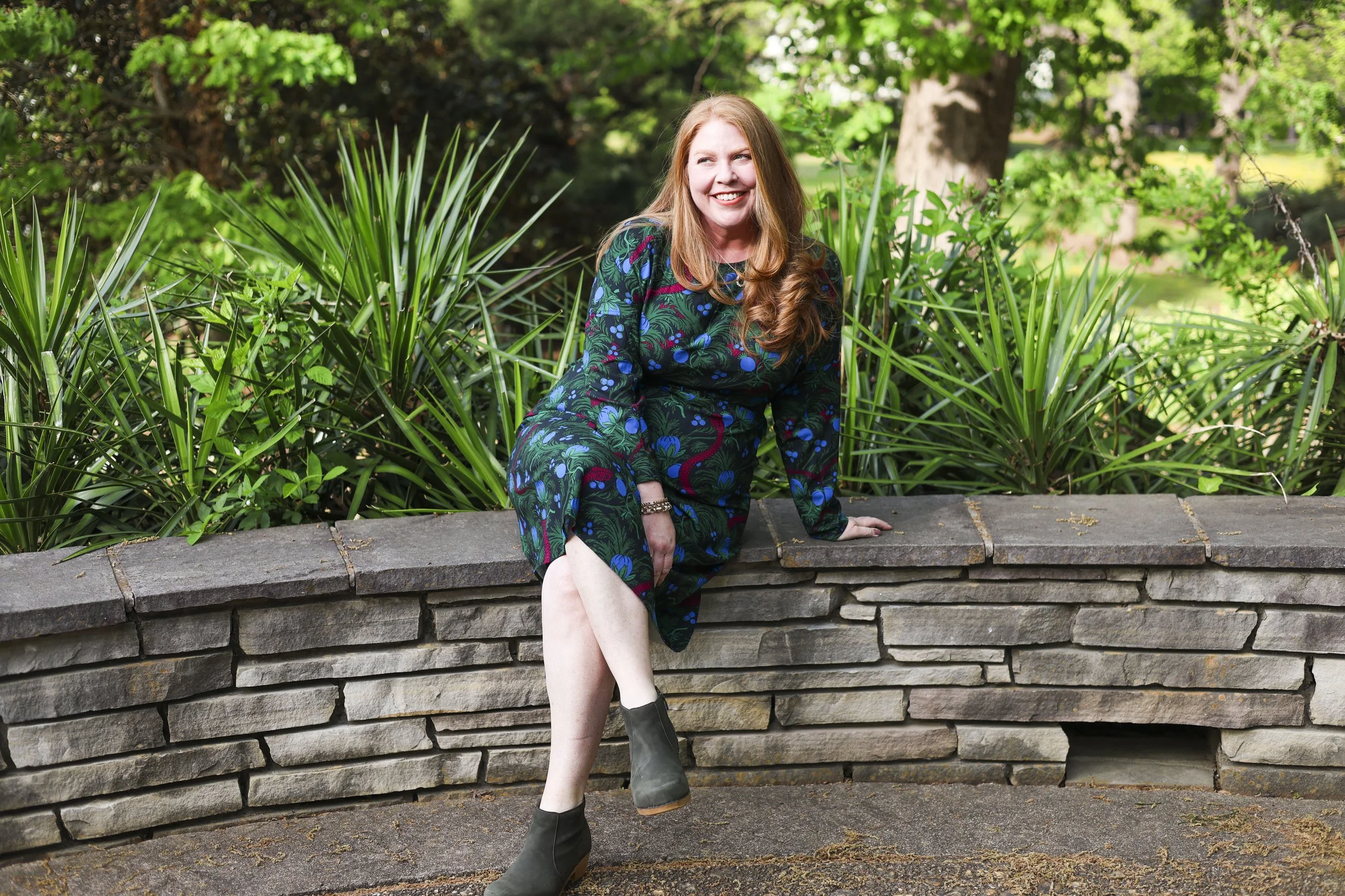 A woman with red hair wearing a dark floral dress and green boots sitting on a stone ledge in a park with green trees and plants surrounding her, smiling and looking to her right.