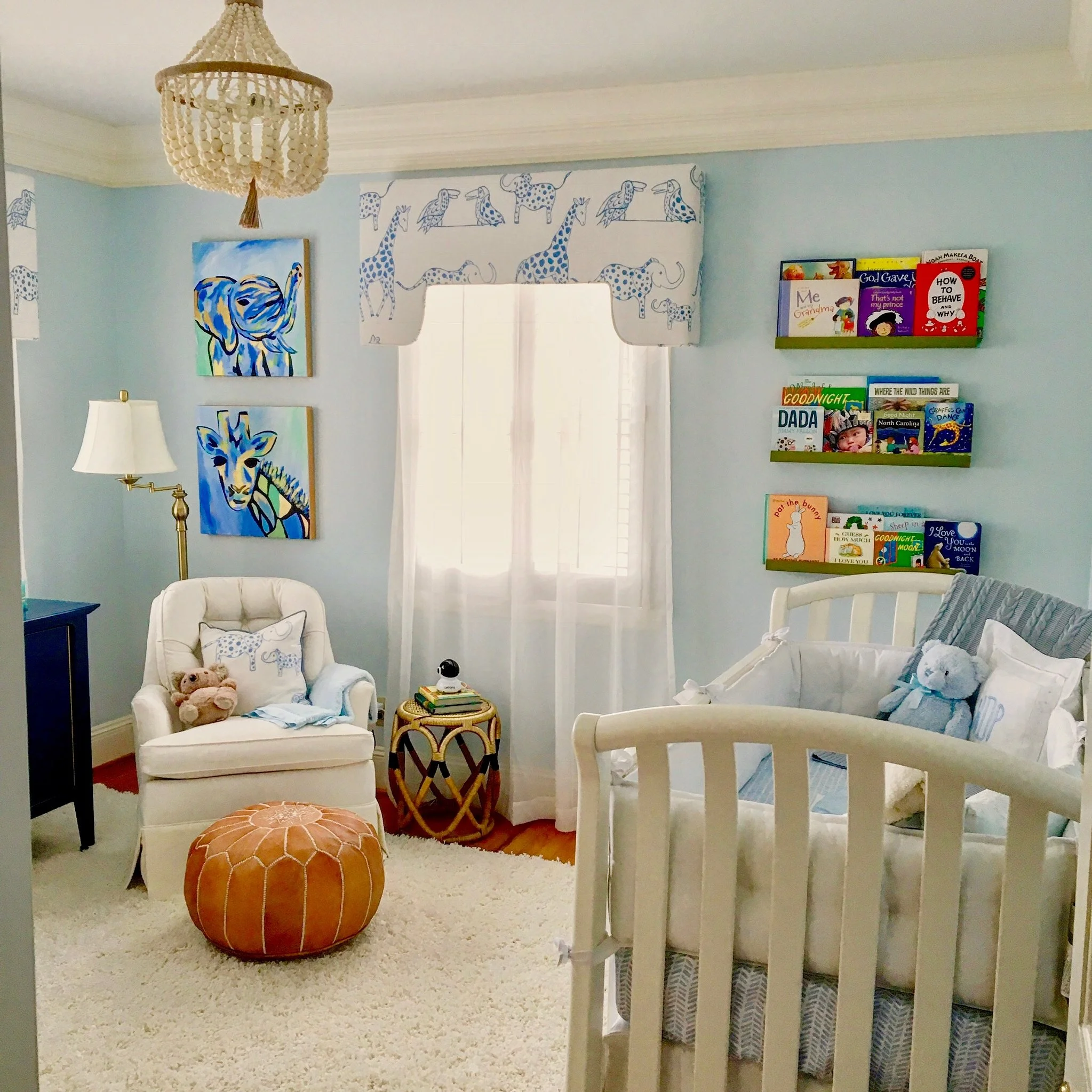 A cozy children's bedroom decorated with blue walls, featuring animal-themed artwork, a bookshelf filled with children's books, white furniture, a beaded chandelier, and plush toys.