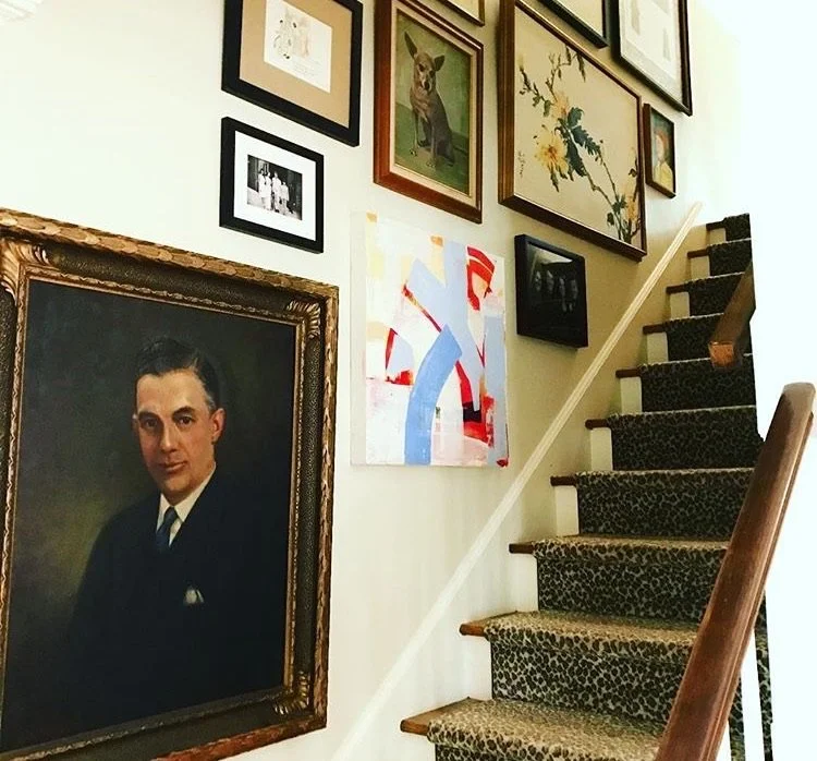 A staircase with a leopard print carpet runner and a wooden handrail, with multiple framed artworks and photographs on the wall beside it, including a large portrait of a man in a suit.