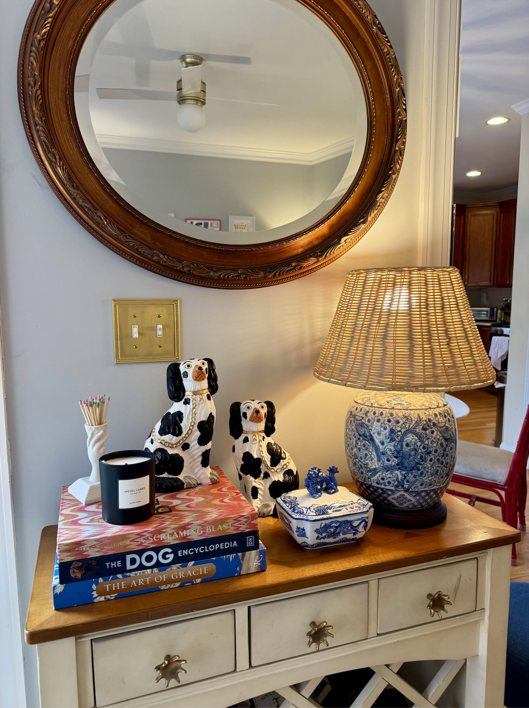 A sideboard with a wooden top decorated with two black and white dog ceramic figurines, books titled "Scrambling Blast," "The Dog Encyclopedia," and "The Art of Gracie," a black candle, a white ceramic container with swabs, a blue and white patterned table lamp, a blue and white porcelain box with small dog figurines, and a large round mirror with a wooden frame hanging on the wall behind.
