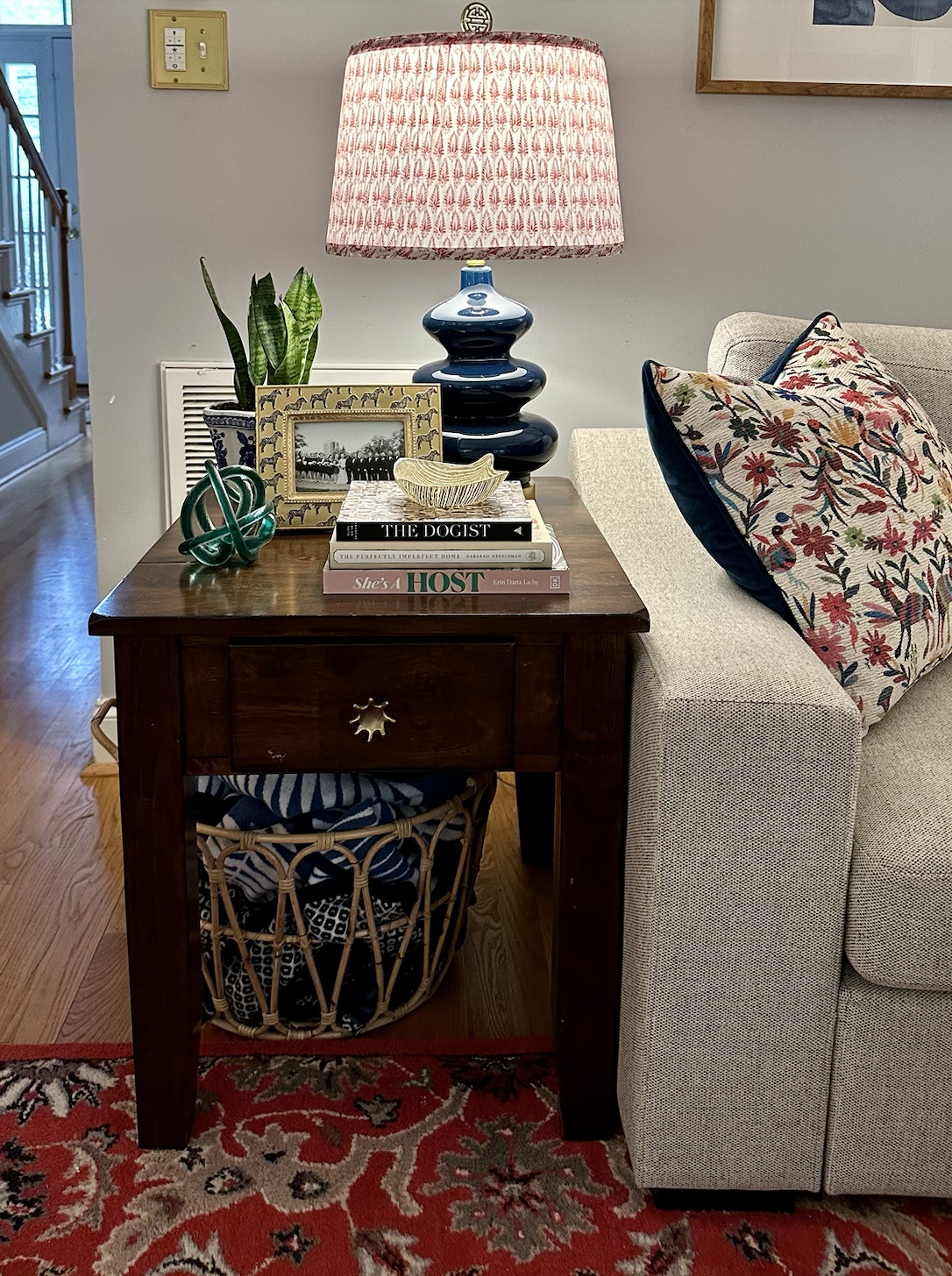 A living room corner with a beige sofa and a wooden side table. The table holds a blue and white ceramic lamp with a red patterned lampshade, a potted snake plant, a decorative green knot sculpture, framed black and white photo, and books. A wicker basket with blankets is underneath the table. The floor has a red patterned rug, and a staircase is visible in the background.