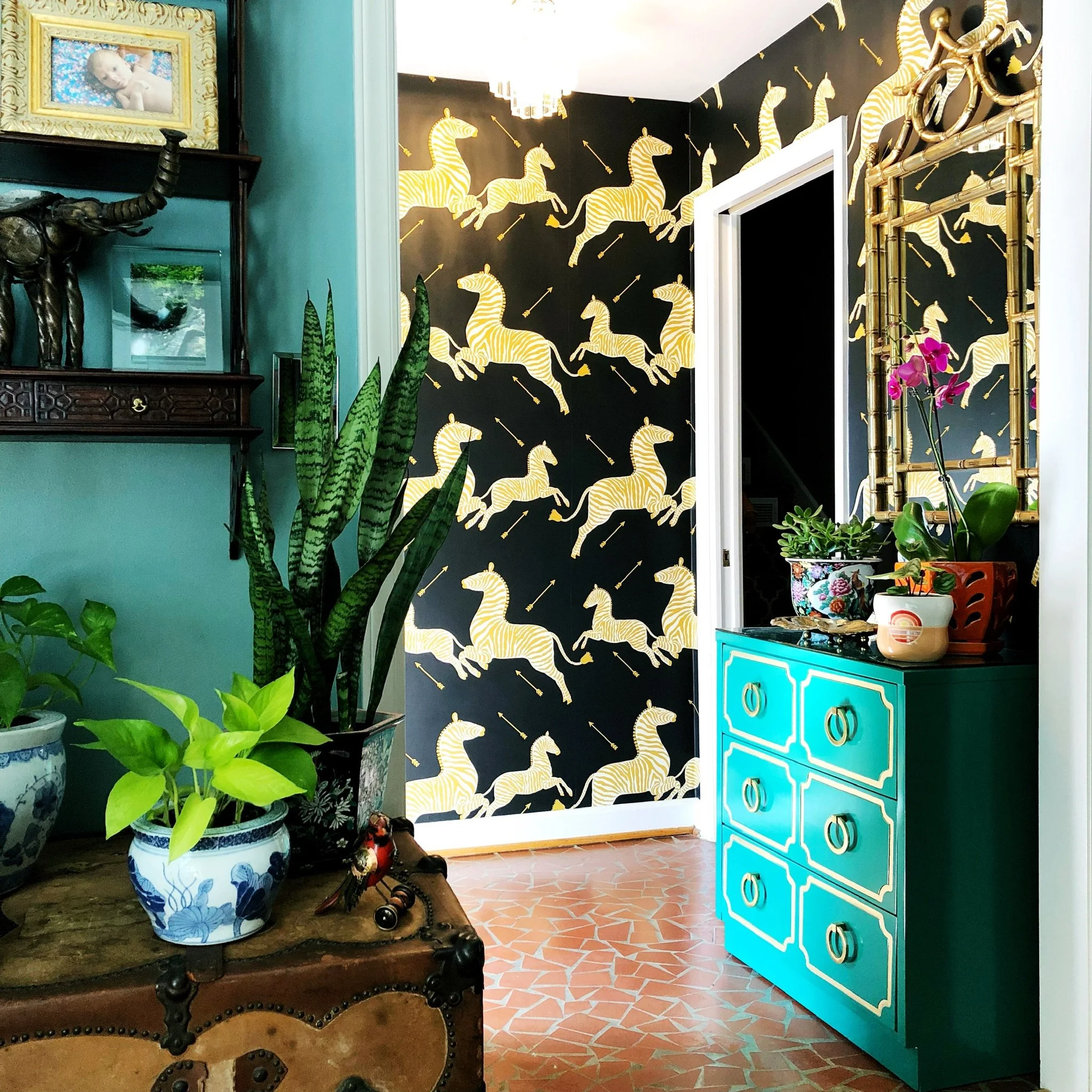 Interior of a room with a black wall decorated with yellow zebras and arrows. A teal dresser with gold accents is visible on the right. There are green plants in decorative pots, an ornate wooden chair, and framed artwork. A gold mirror frame and a floral container are also part of the decor.