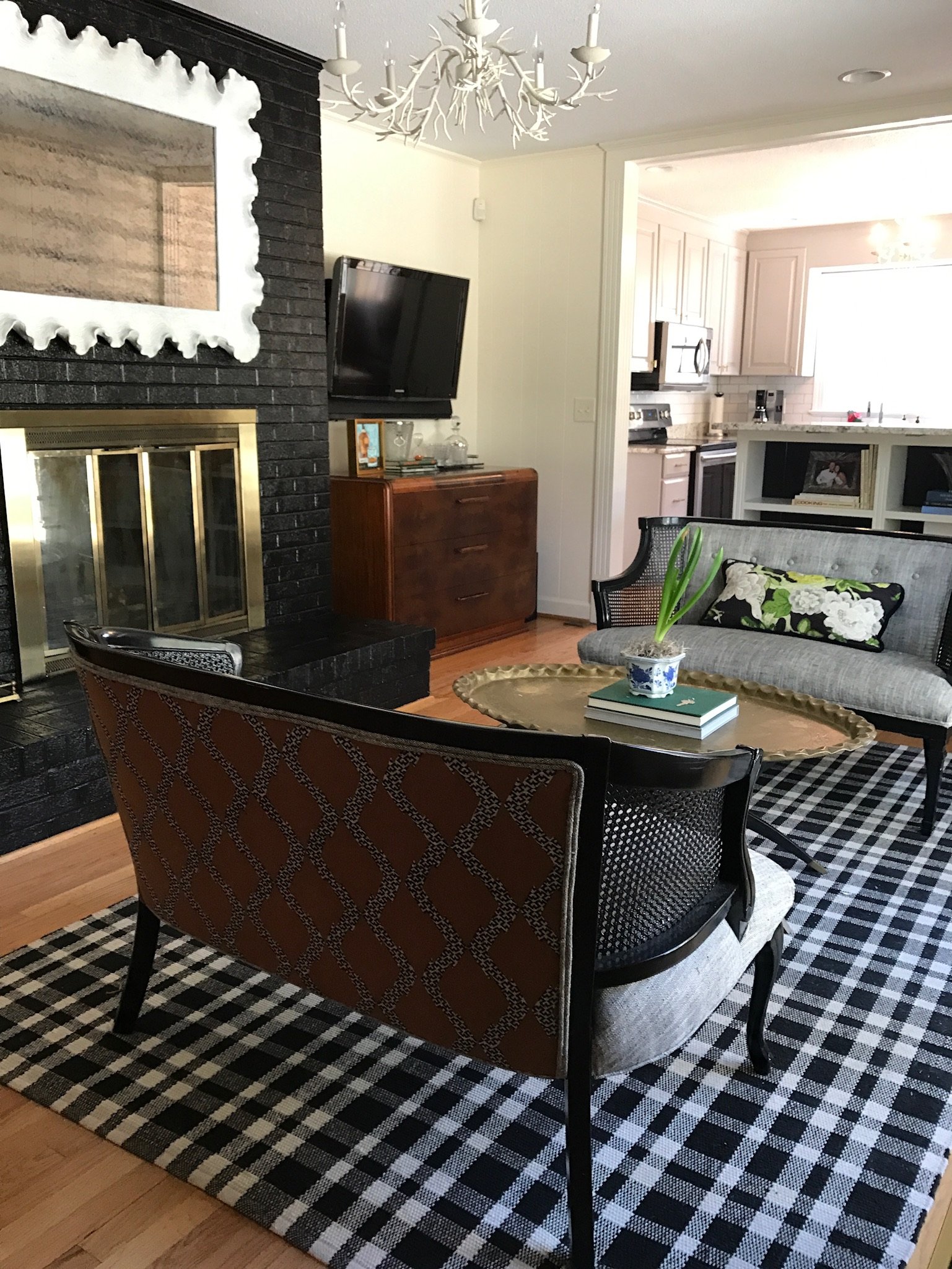 Living room with black brick fireplace, a wooden dresser with a TV, and a white kitchen in the background. There is a black wicker chair, a patterned black and white rug, and a gray sofa with a floral pillow. A potted plant is on a gold tray on the coffee table, and a chandelier with antler-like arms hangs from the ceiling.