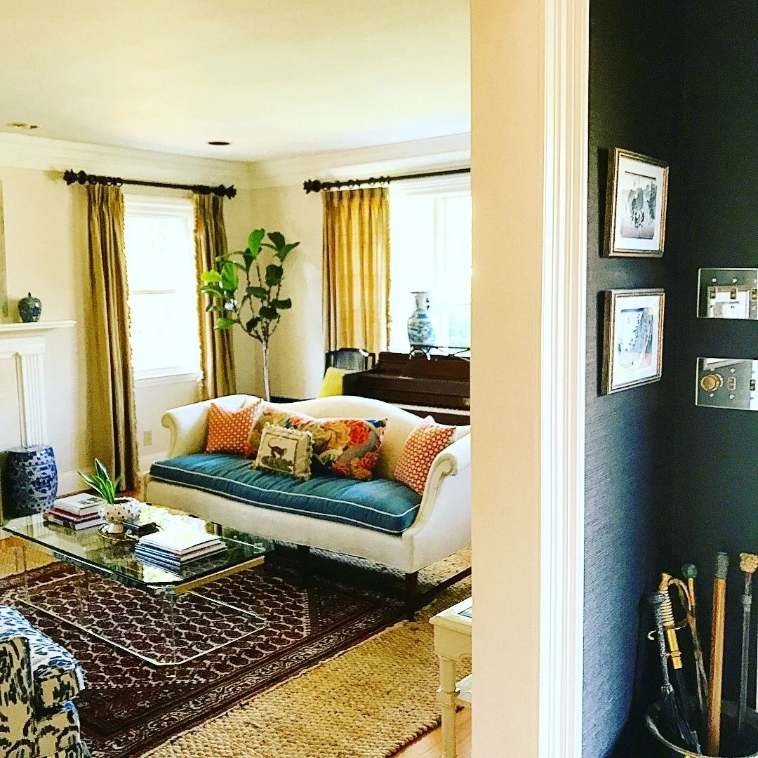 Living room with yellow curtains, white couch with colorful pillows, a glass coffee table, a potted plant, and a dark wall with framed pictures.