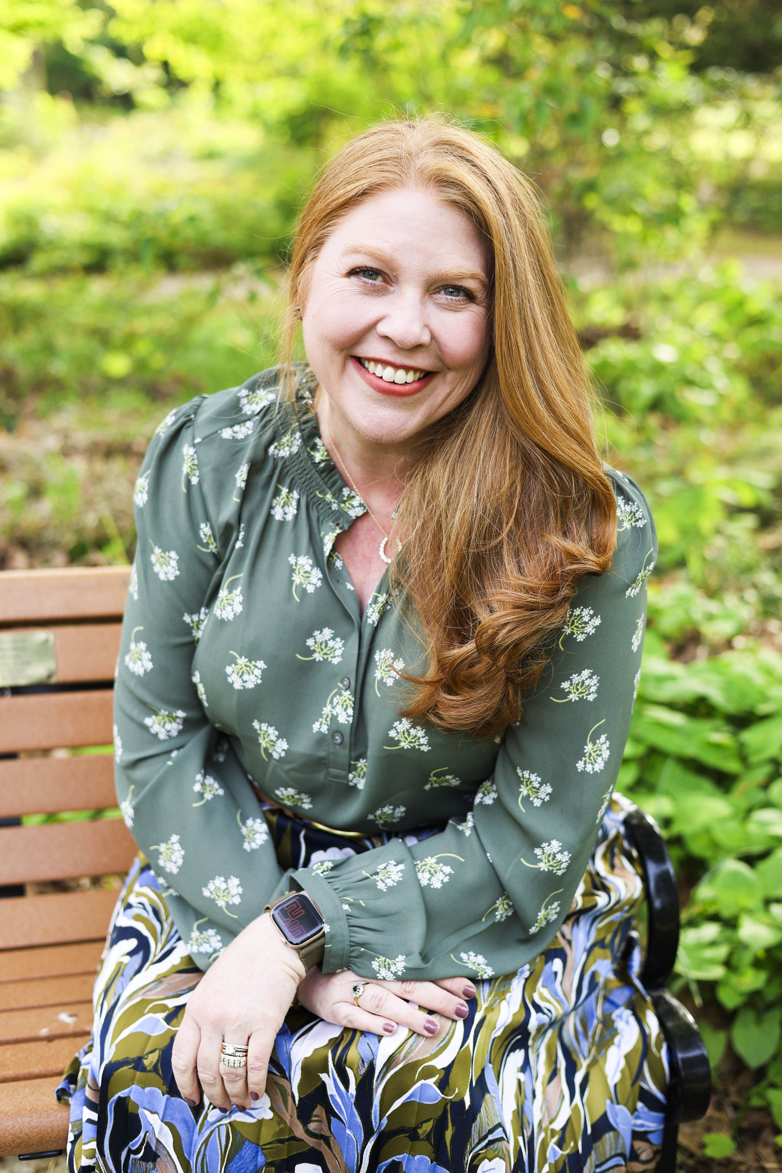 A smiling woman with long red hair, green eyes, and light skin sitting outdoors on a wooden bench surrounded by greenery, wearing a green floral blouse, patterned skirt, and a smartwatch.
