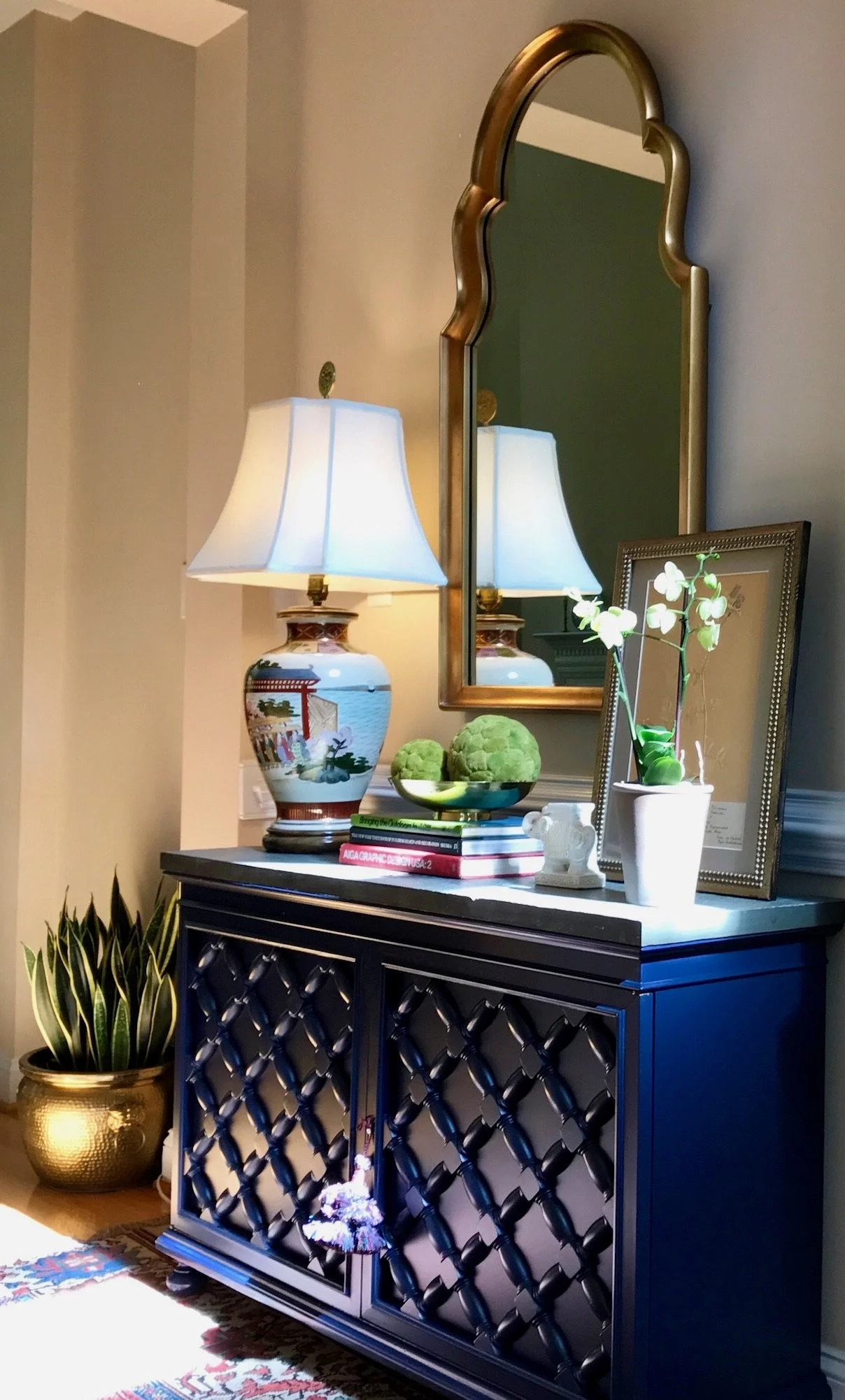Decorative console table with a lamp, a framed picture, a white orchid plant, and a bowl of green artichokes. A large mirror hangs above, reflecting the lamp. To the left, a potted snake plant with variegated leaves in a gold pot. The table has a black lattice design on its front.