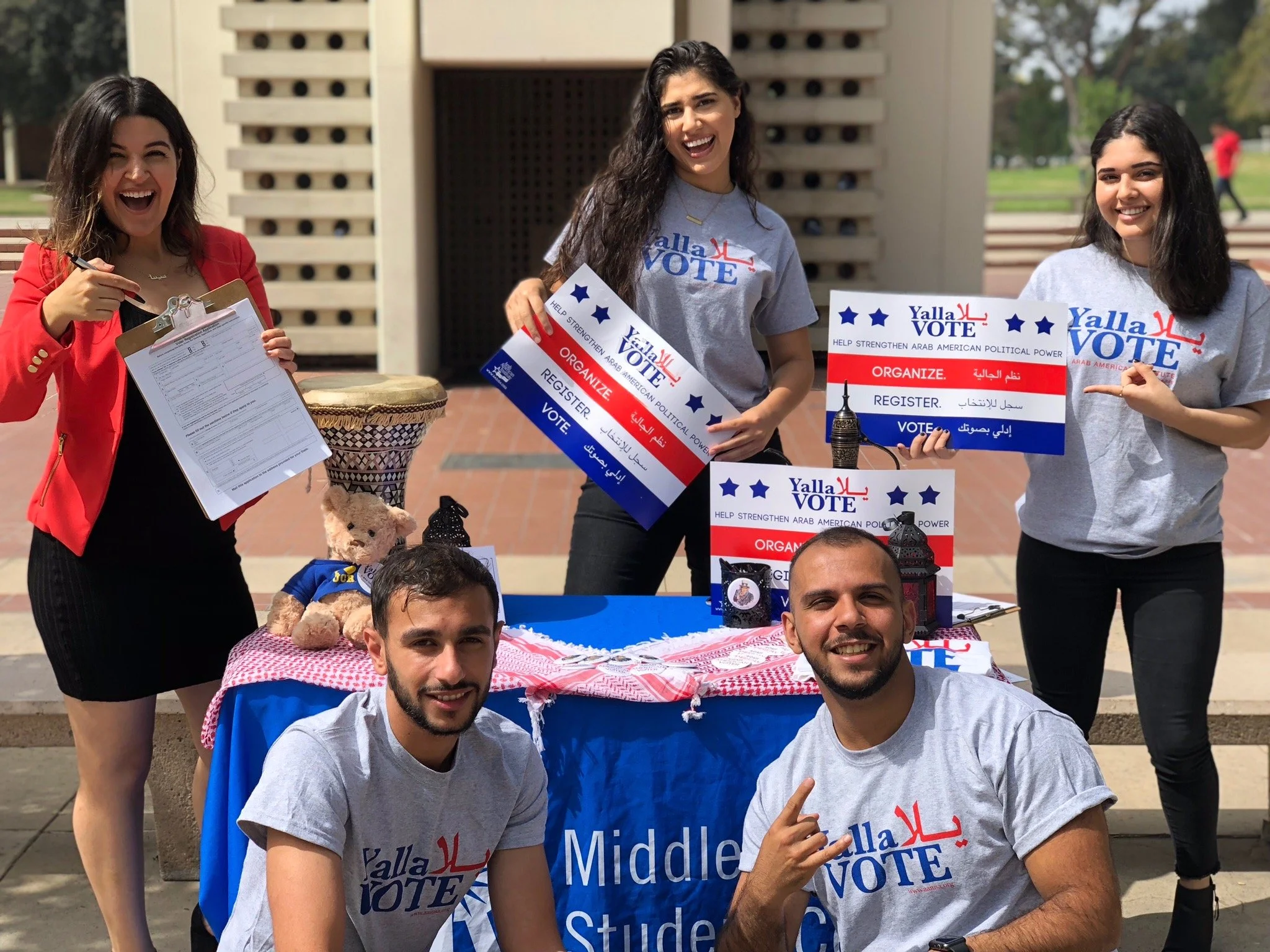 Group of young adults at a voting campaign booth, holding signs and materials promoting Arab American political participation, outdoors on a sunny day.