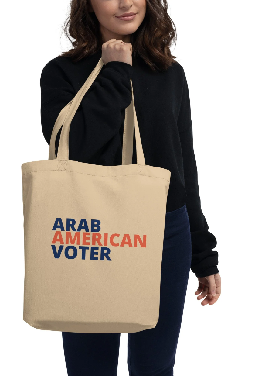 A woman holding a beige tote bag with the words 'ARAB AMERICAN VOTER' printed in blue and red, while wearing a black long-sleeve shirt and dark blue jeans.