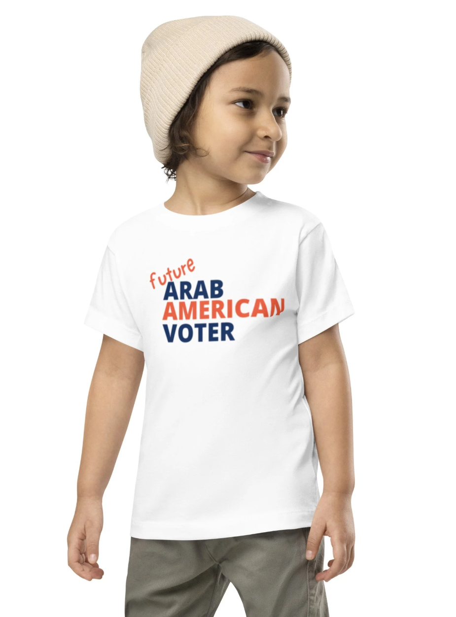 A young boy wearing a beige knit cap and a white T-shirt with the words 'future ARAB AMERICAN VOTER' printed in bold, colorful letters.