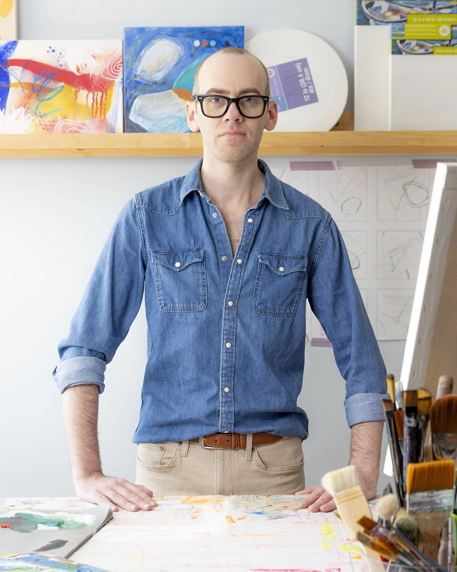 The artist, a white man in glasses and a denim shirt, stands behind an art table with his hands on the surface. in an art studio with colorful paintings and art supplies around him.