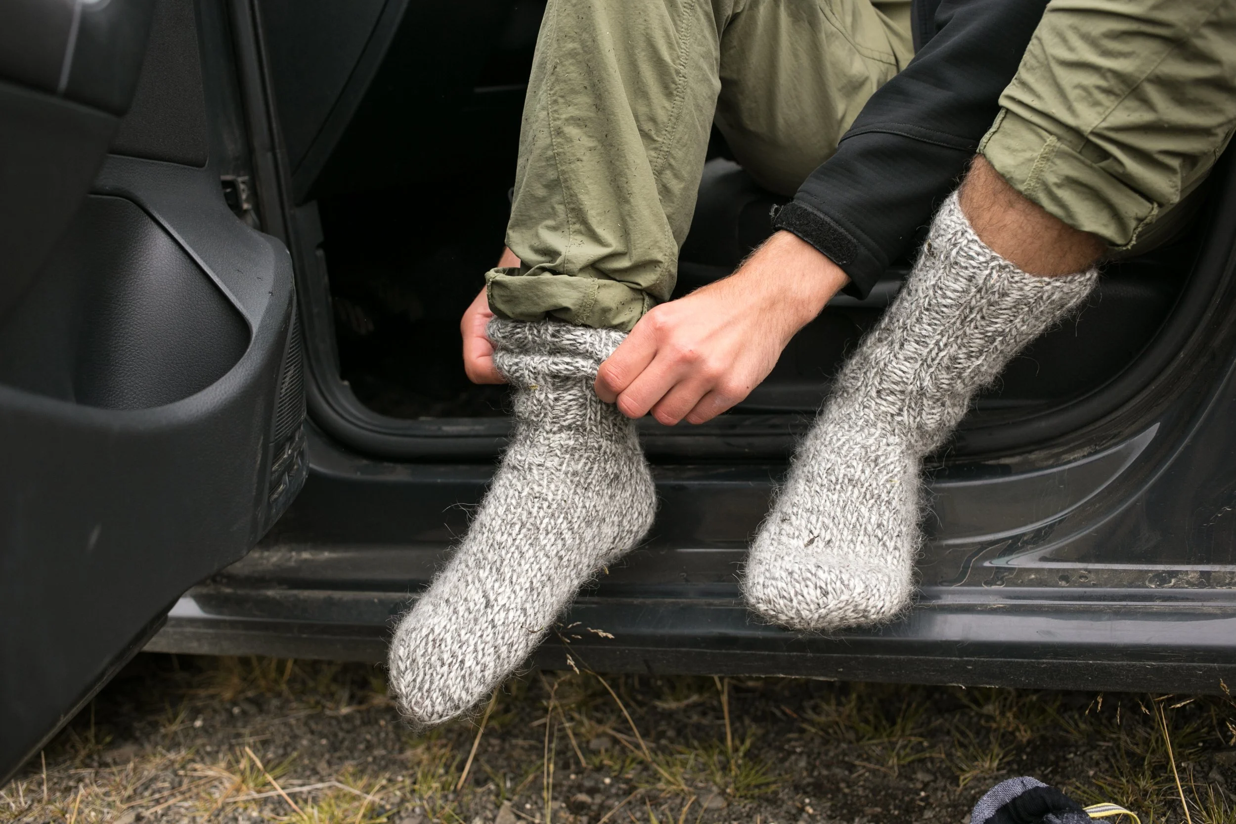 A man has his car door open and is pulling thick, woolen socks onto his feet.