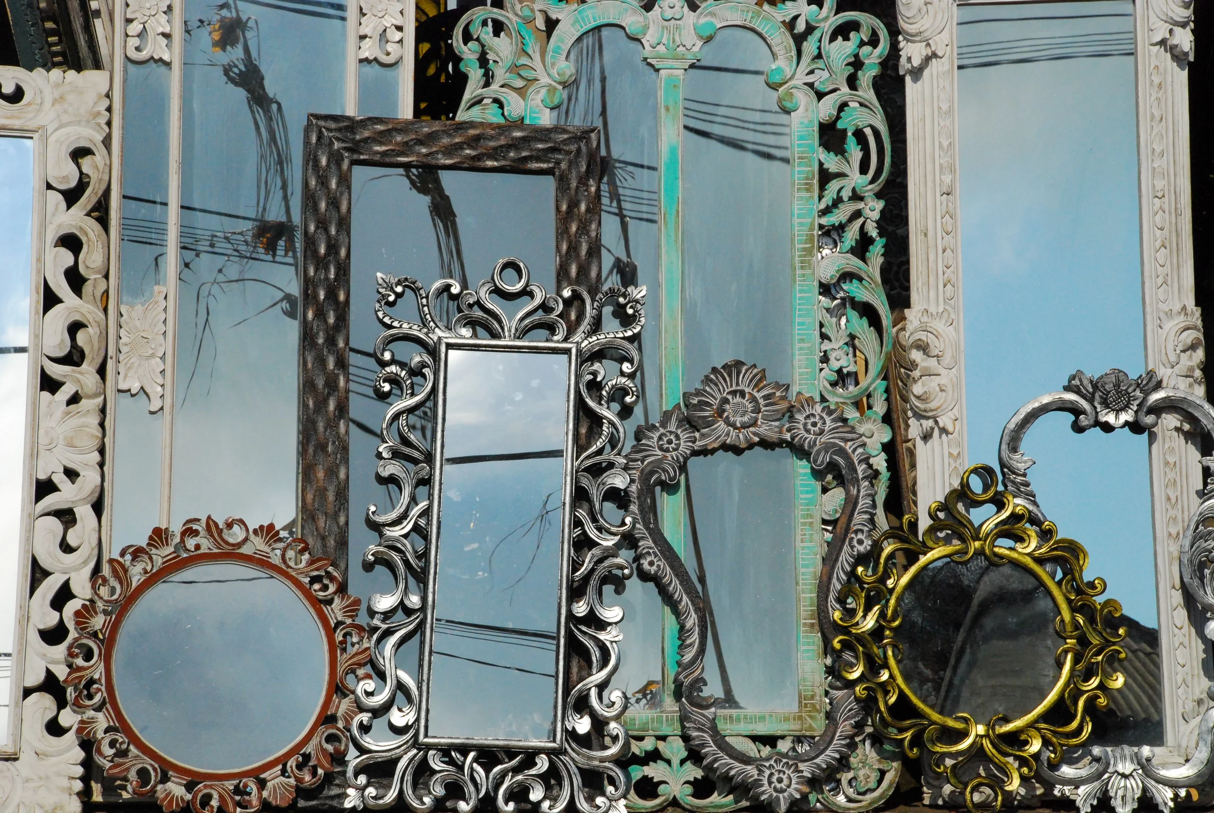 A collection of antique mirrors, large and small, lean against a building and reflect blue sky and power lines.