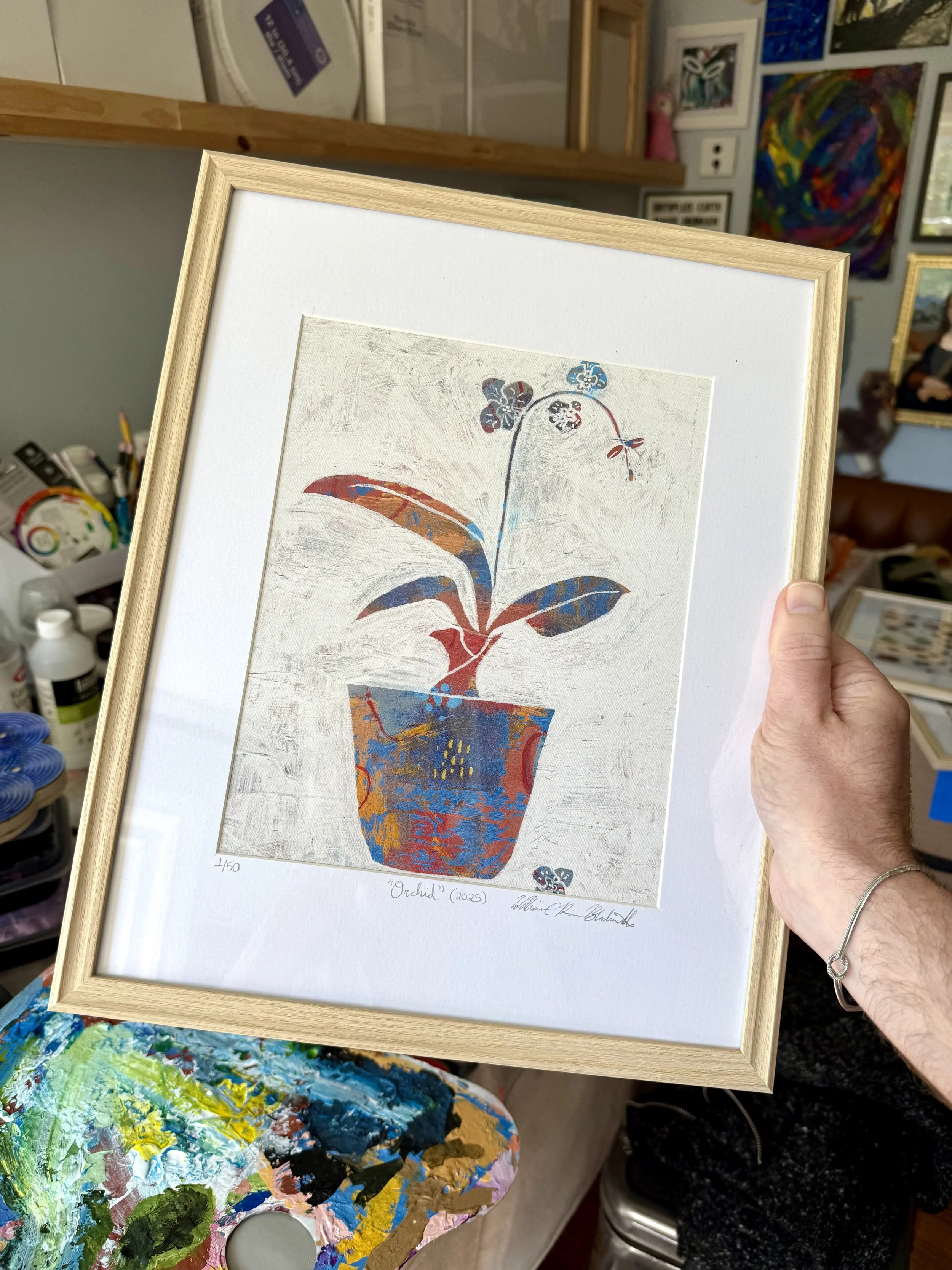 A framed print of my "Orchid" (2025) painting in reds, ochres, and blues, surrounded by white matting that is signed, numbered, and titled in graphite, all in a light wooden frame. My worktable in my studio can be seen behind it.