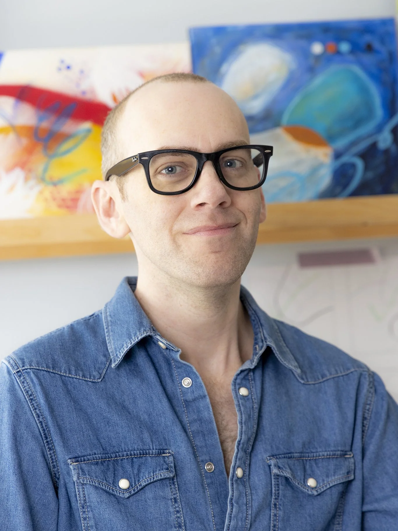 A man with short, light hair wearing black glasses and a blue denim shirt, standing in front of colorful abstract paintings.