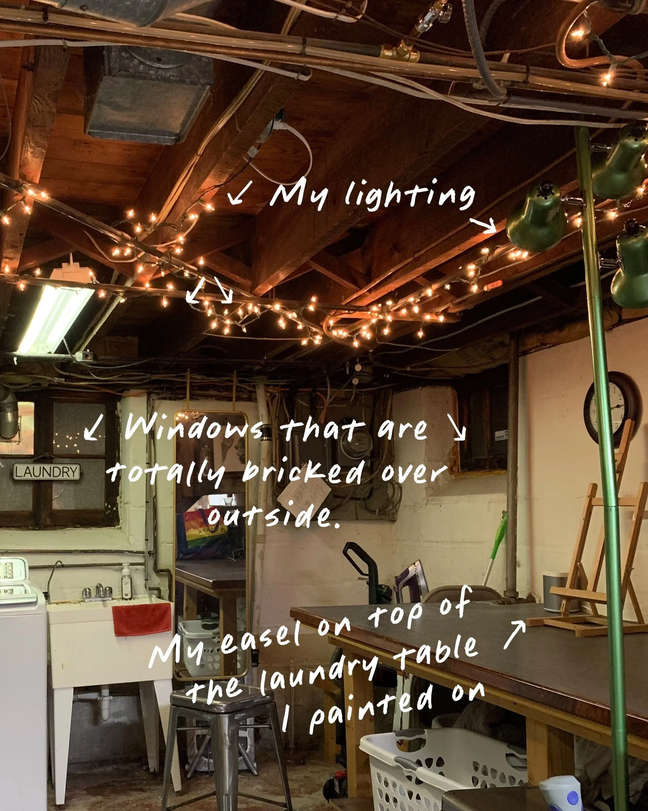 Unfinished basement workspace: string lights overhead, bricked-over windows, and an easel balanced on the laundry table—a self-aware look at creative compromise.