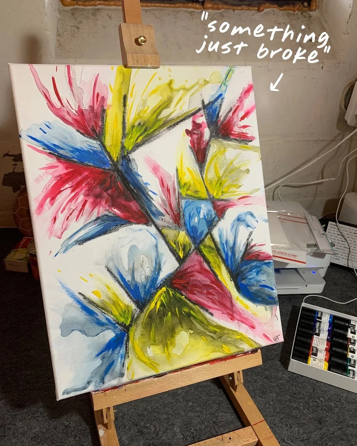 Painting titled 'something just broke'—abstract almost-floral shapes in bold reds, yellows, and blues burst out of dark black lines on the white canvas, displayed on a basement studio easel.