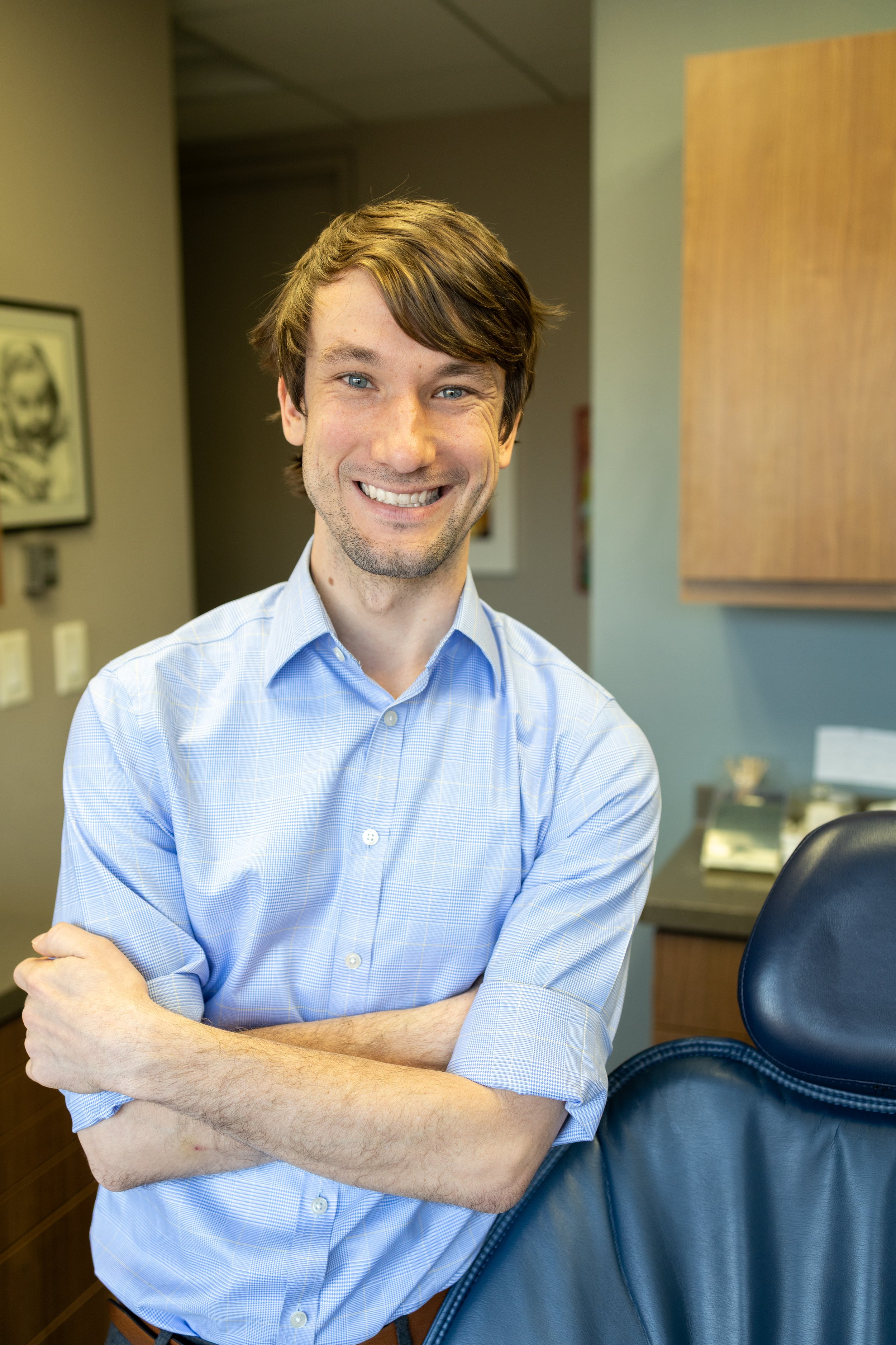 A smiling man with brown hair, blue eyes, and a blue collared shirt leaning against a dental exam chair