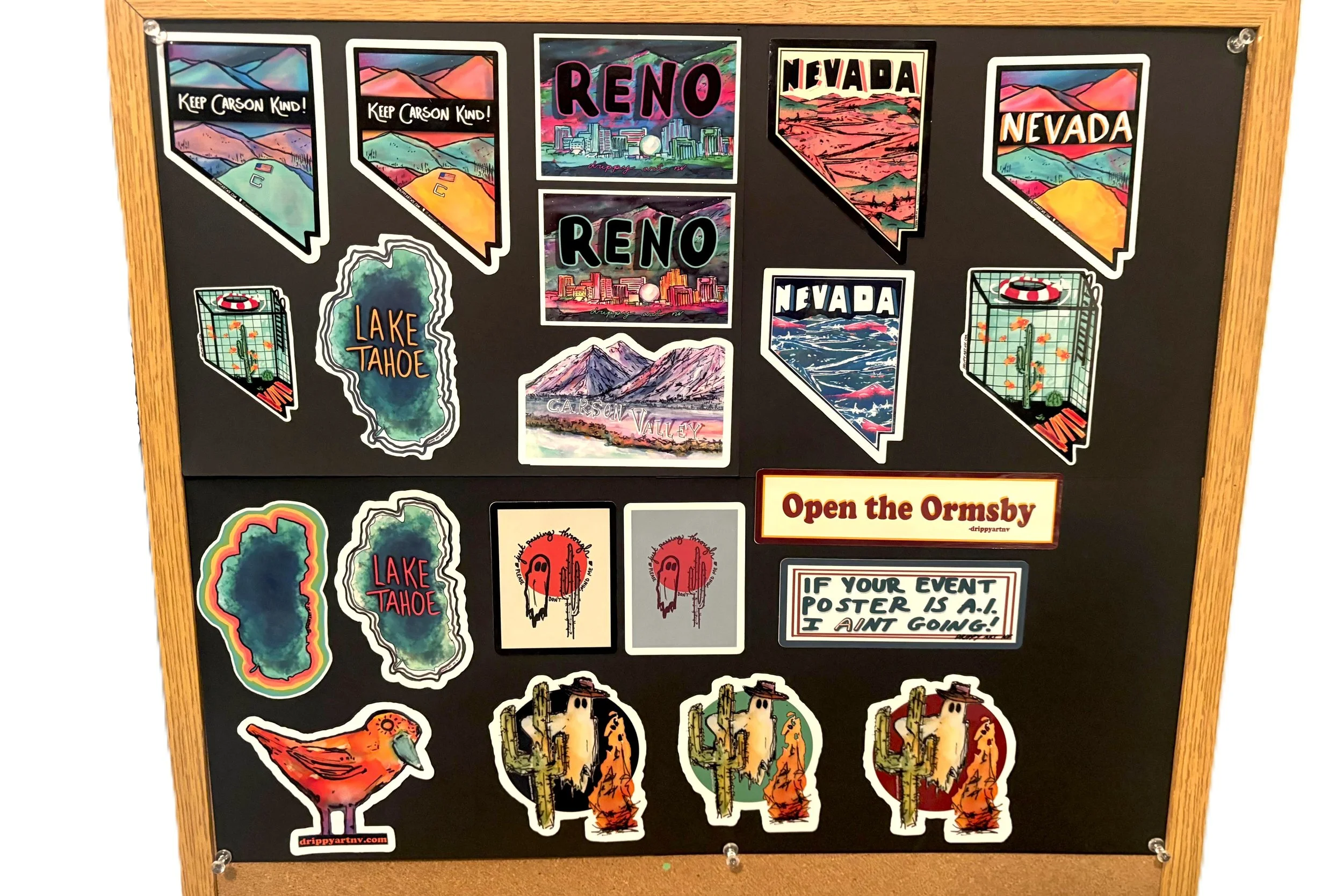 A bulletin board with various colorful stickers and artwork. The stickers include images of lakes, mountains, cacti, and birds. Some stickers have text such as "Keep Carson Kind!", "Reno", "Nevada", "Lake Tahoe", and "Carson Valley." There is also a sticker saying "Open the Ormsby" and another with the message "If your event poster is a... I ain't going!".