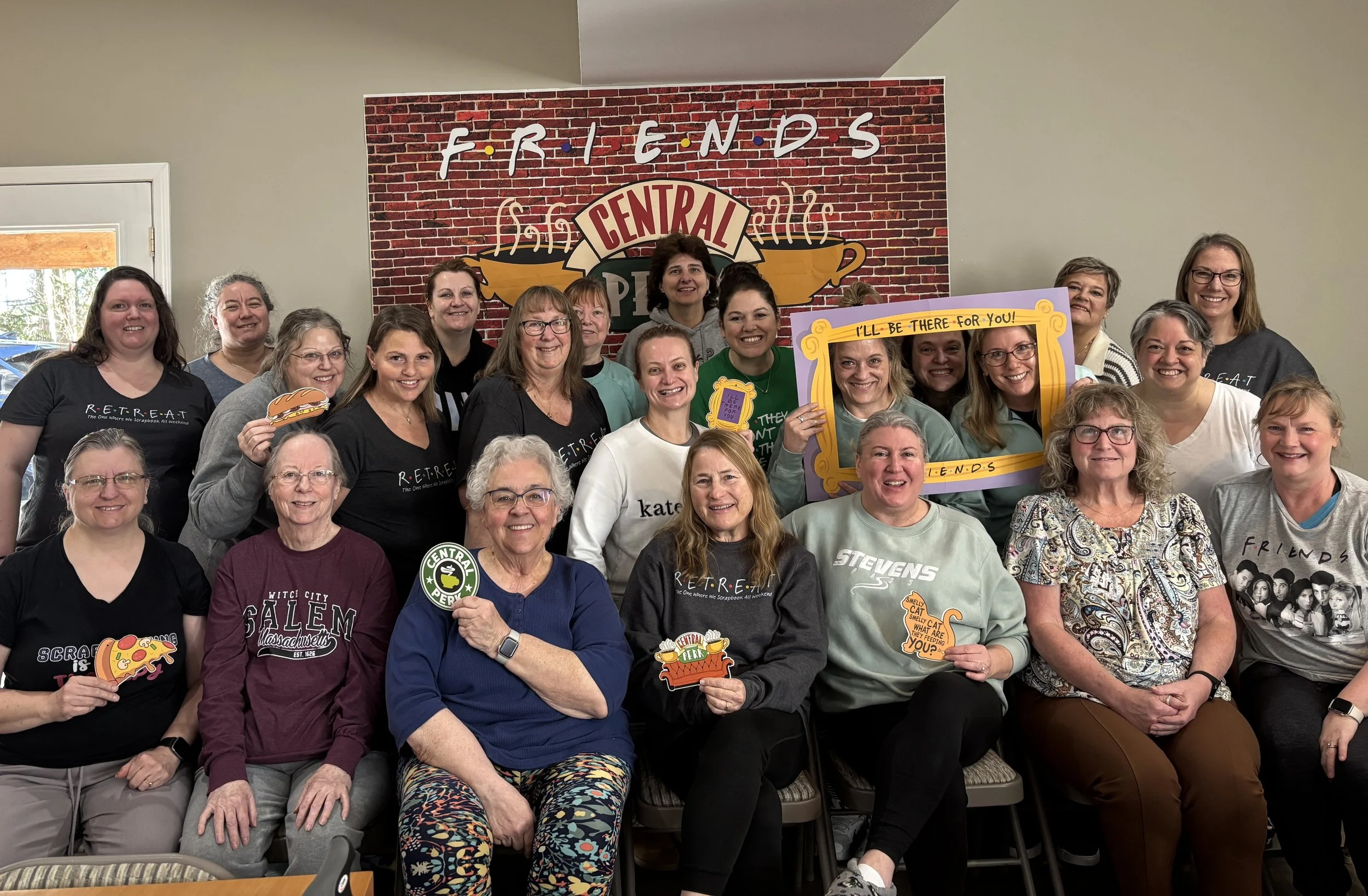 Group of women posing for a photo in front of a wall with a brick background and a sign that reads 'FRENDS'. Many women are smiling and holding various signs and cutouts, some with food-themed designs.