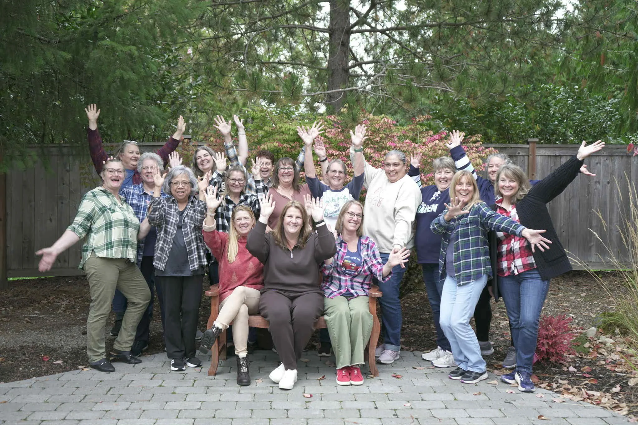 A group of women posing outdoors in front of a wooden fence, with trees and autumn foliage in the background, smiling and waving at the camera.