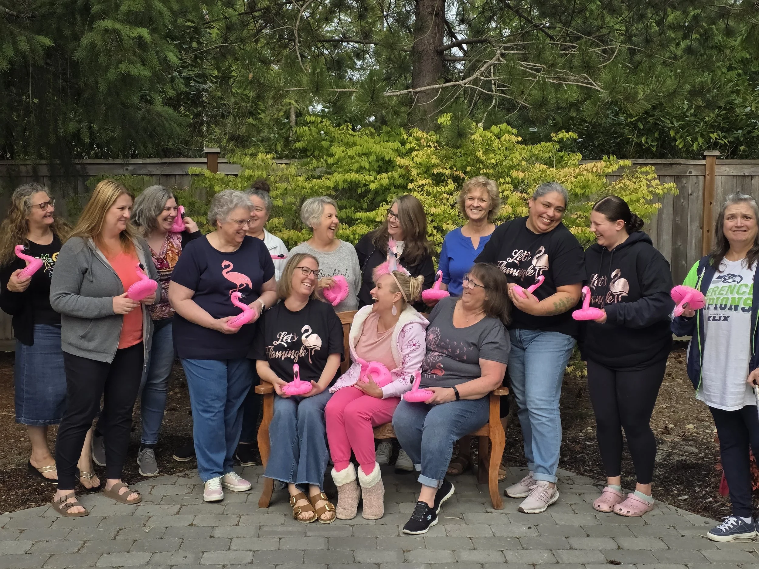 Group of women gathered outdoors, some holding pink flamingo-shaped decorations, smiling and laughing together with a wooden fence, trees, and greenery in the background.