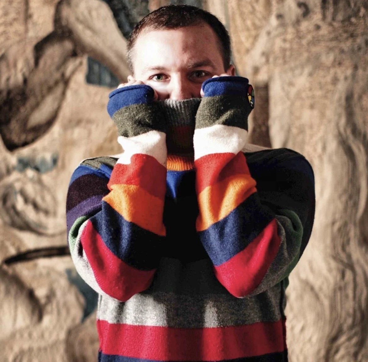 A person wearing a multicolored striped sweater and matching gloves covering their face with fists, standing in front of a textured rock wall.