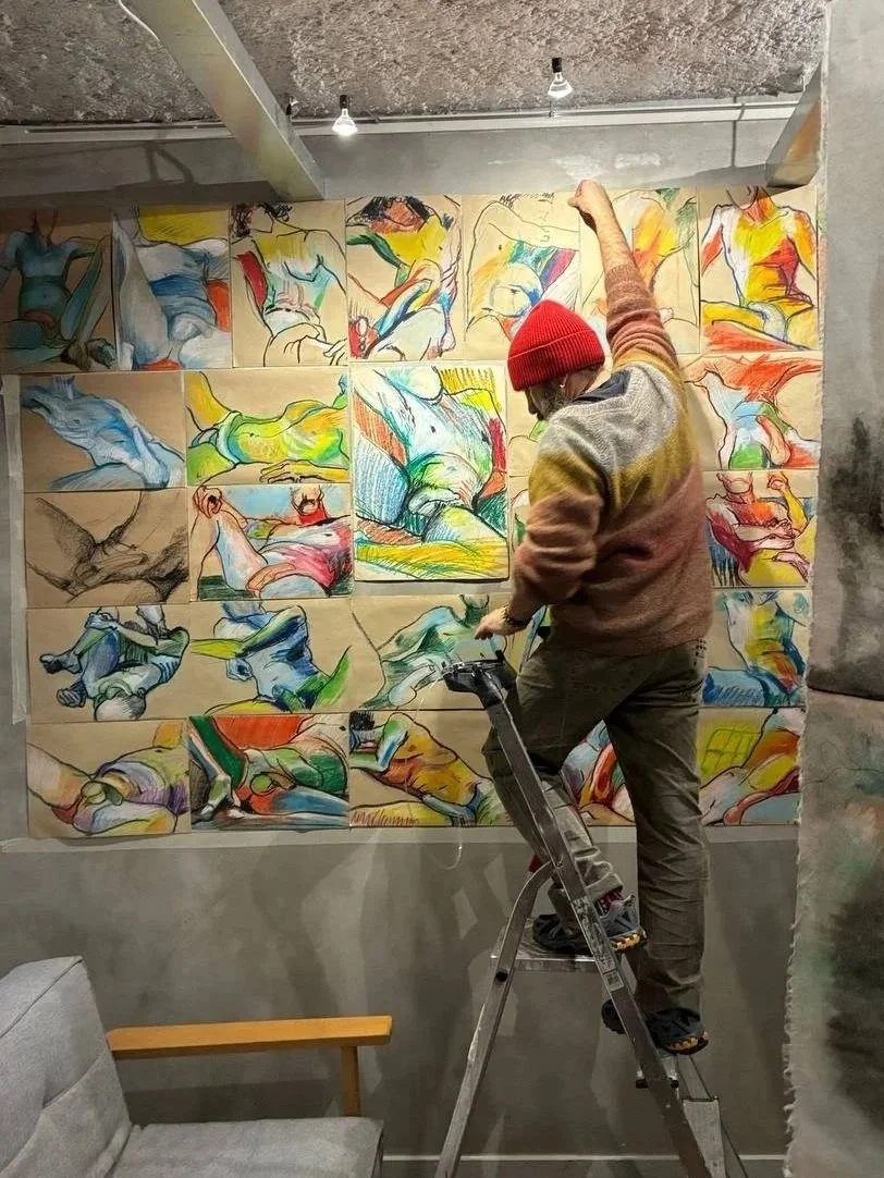 A person wearing a red beanie and colorful sweater is standing on a step ladder, working on a large wall mural made of multiple colorful sketches of human figures in various poses.