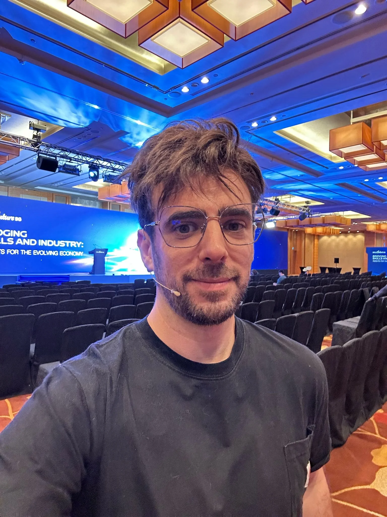 A man with glasses and a goatee taking a selfie in a large conference room with rows of chairs and a stage with a large digital screen