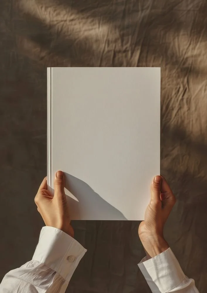 Hands holding a blank white book or notebook with long sleeves, against a textured brown background.