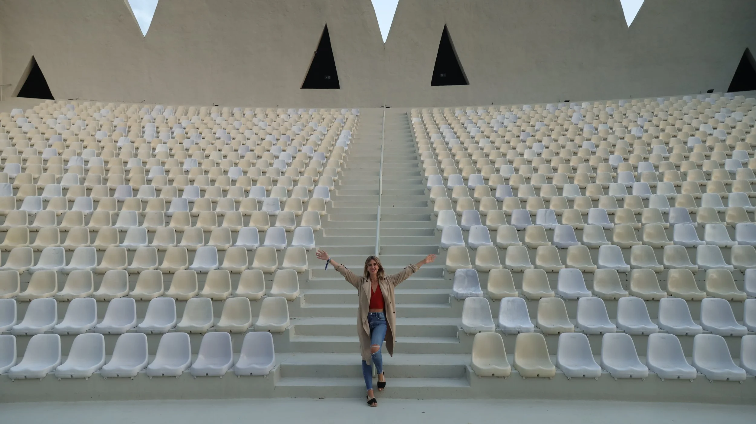 Woman standing on stairs in empty amphitheater with white seats, arms raised, smiling.